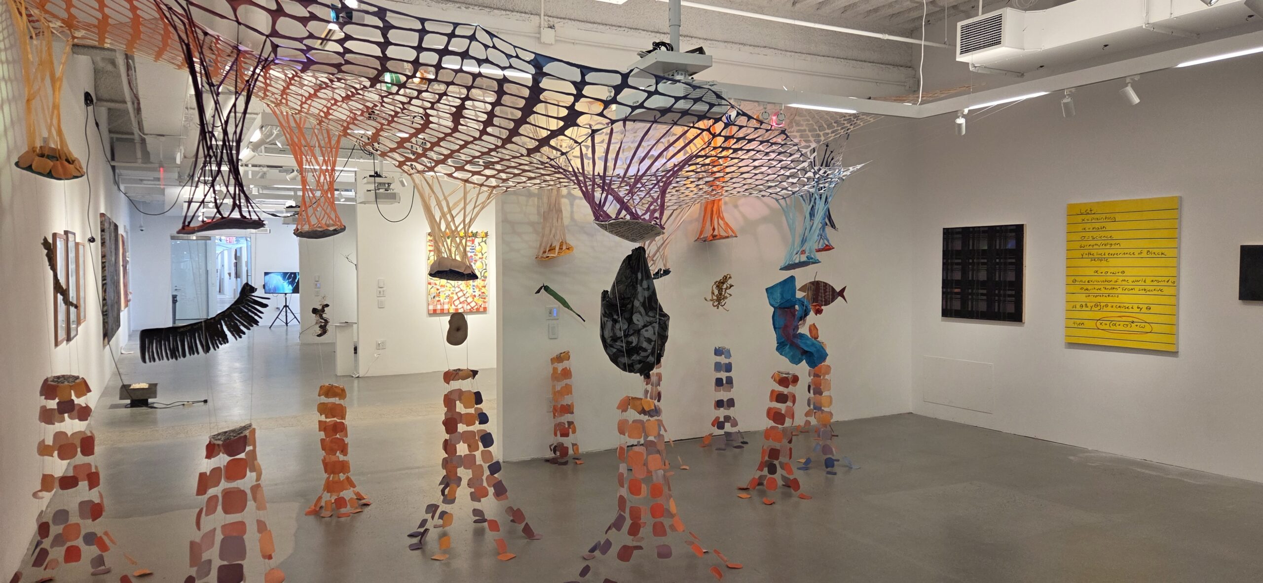 An art gallery interior featuring a series of hanging sculptures made from colorful net-like materials and fabric, resembling organic shapes. The floor has several tall, patterned bases in warm tones. In the background, several framed artworks are hung on white walls, including a bright yellow piece with handwritten notes. The lighting is bright, highlighting the textures and colors of the installation.