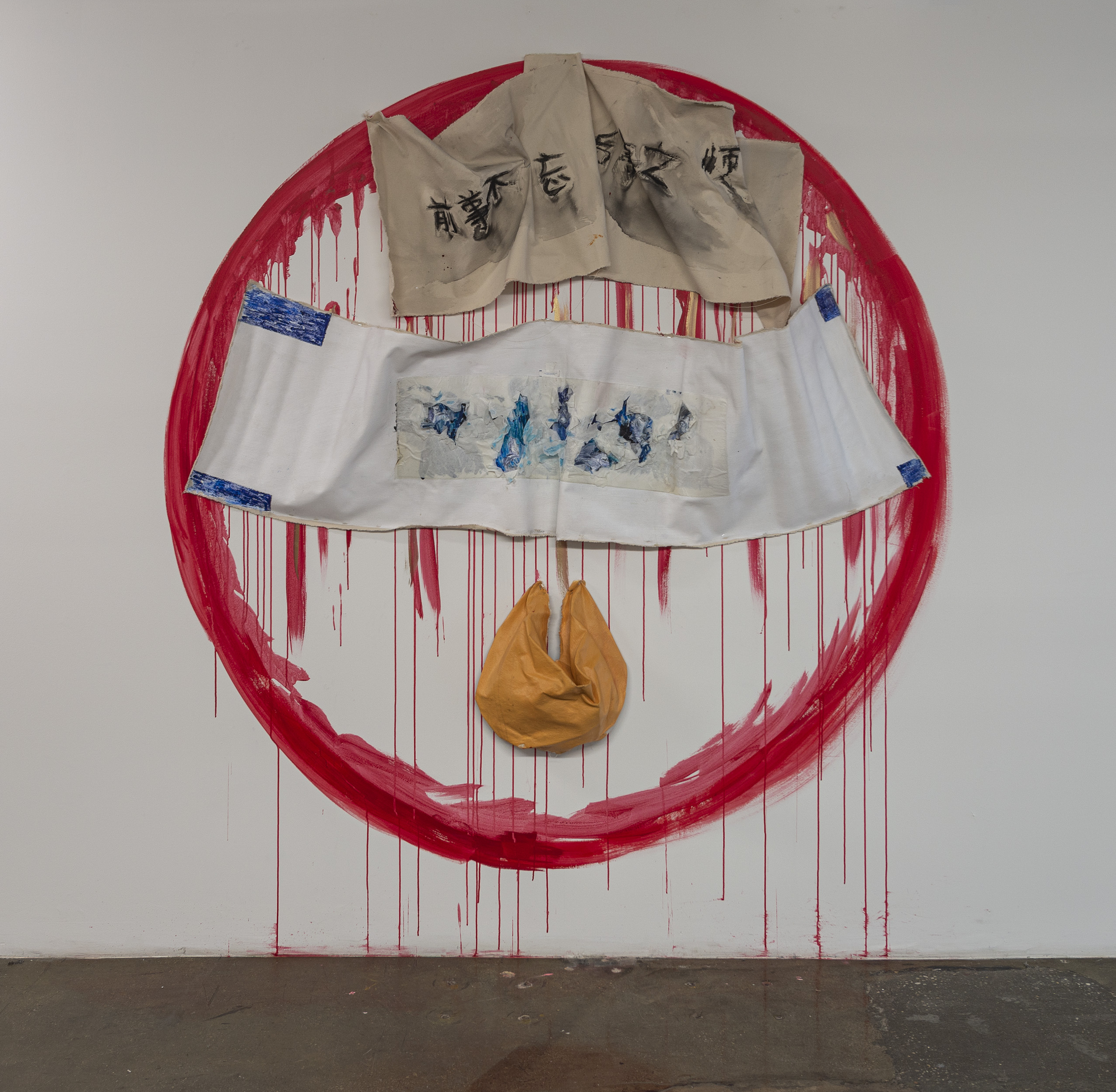 Abstract artwork featuring a large red circular shape on a white wall. At the top, two beige fabric pieces with black script hang. Below them, a horizontal white textile with blue accents and an abstract blue painting is displayed. At the center, a yellow pouch hangs down, with red paint drips creating a contrast against the white background. The overall composition conveys a sense of layered textures and colors.