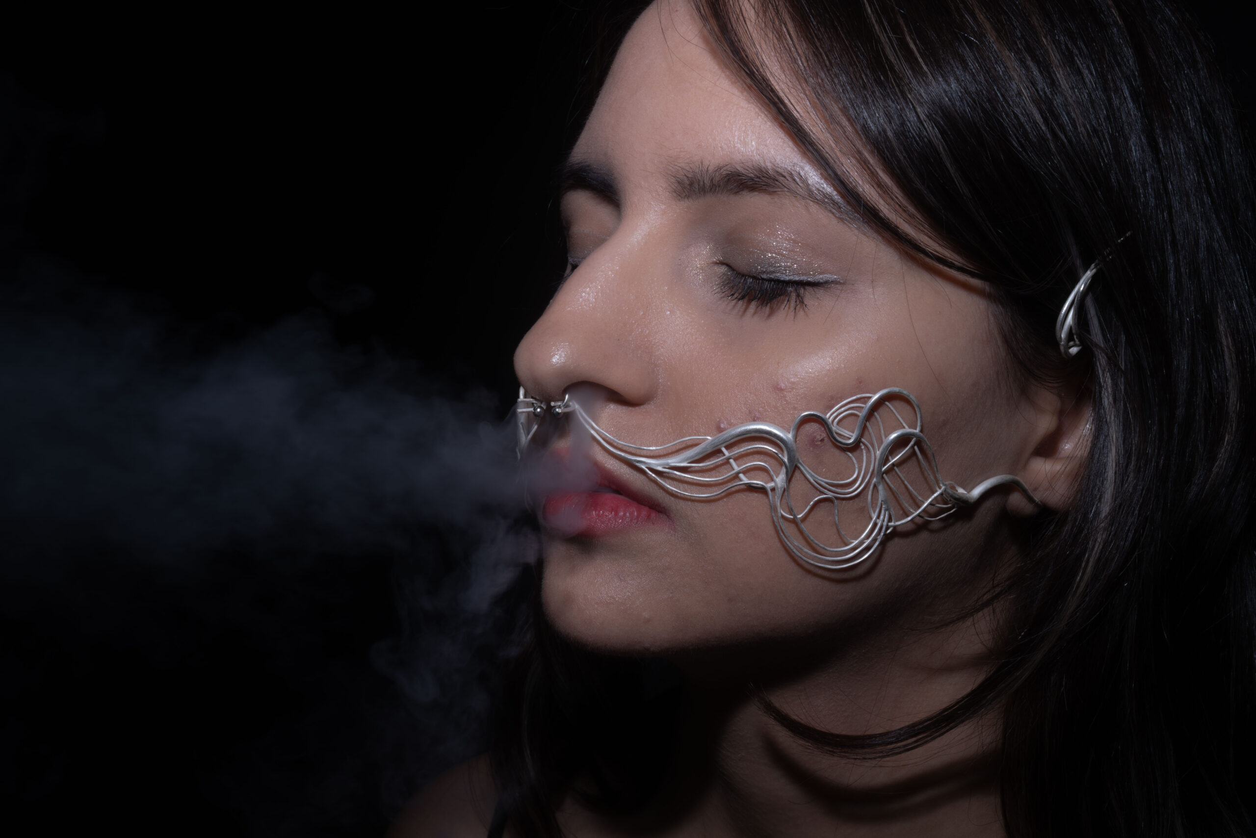 A close-up of a woman's face with her eyes closed, featuring intricate metallic wire designs resembling a mustache around her mouth. She is exhaling vapor, creating a smoky effect in front of her face. The background is dark, emphasizing the details of the wire structure and her features.