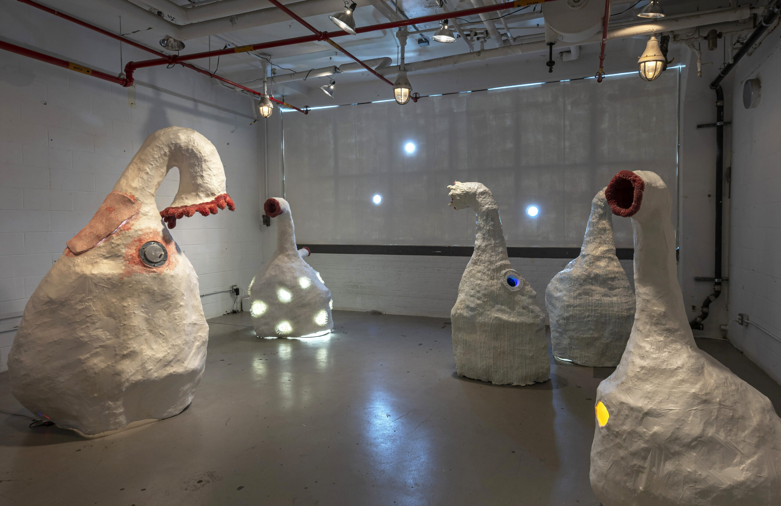 The image depicts an art installation featuring five large, abstract sculptures that resemble bulbous figures with elongated necks. The sculptures are textured and painted in white with colorful accents, including red and blue spots. Some have circular lights integrated into them. The setting is an industrial style room with a white wall, exposed pipes, and overhead lighting. Natural light filters through a window with small circular cutouts.