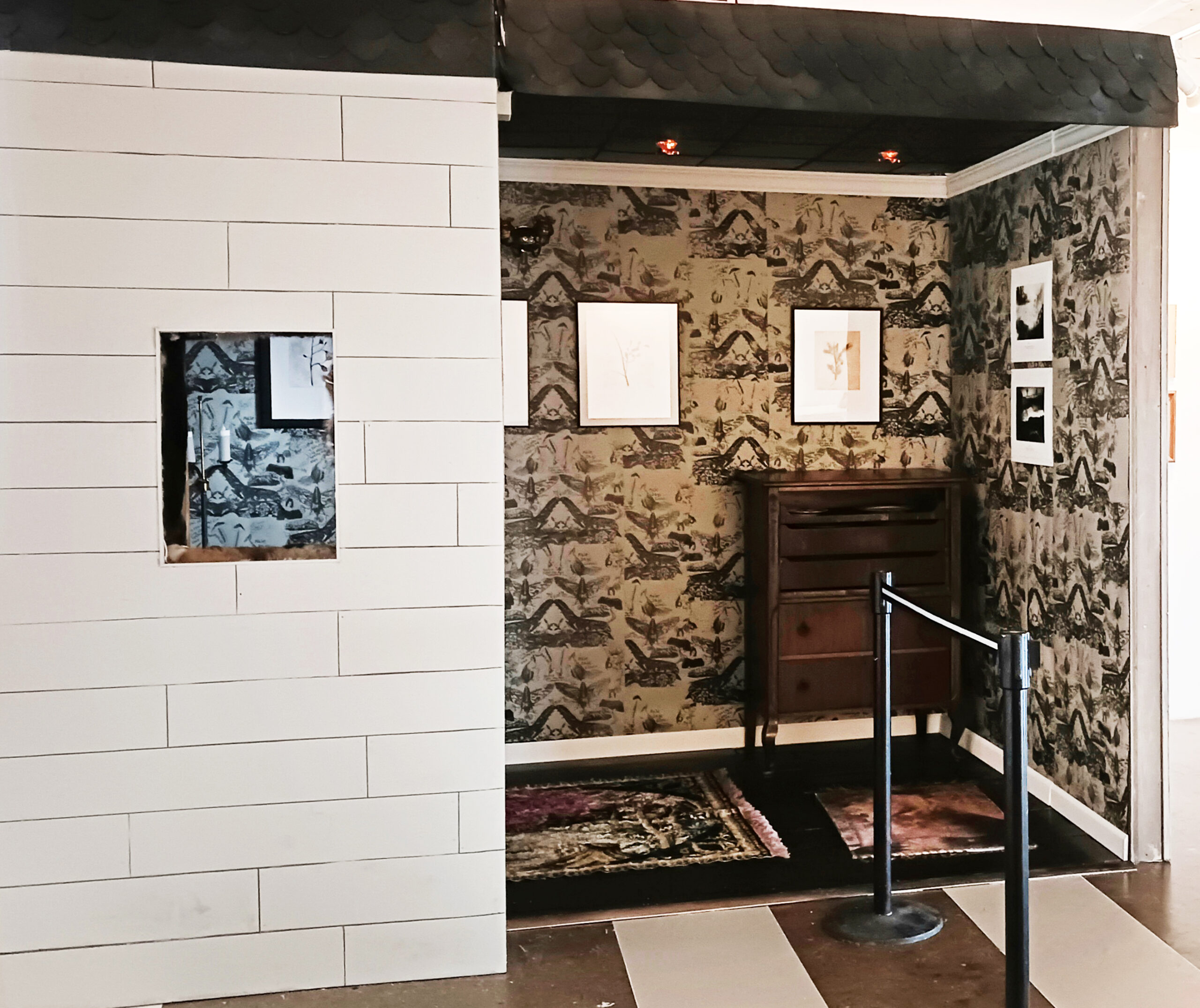 A partially open room features a white wall with a rectangular cutout on the left side. Behind the cutout is a patterned wallpaper with framed artworks displayed. To the right, there is a wooden dresser with multiple drawers and a section of a rug visible on the floor. A retractable barrier is set up to guide visitors in the space.