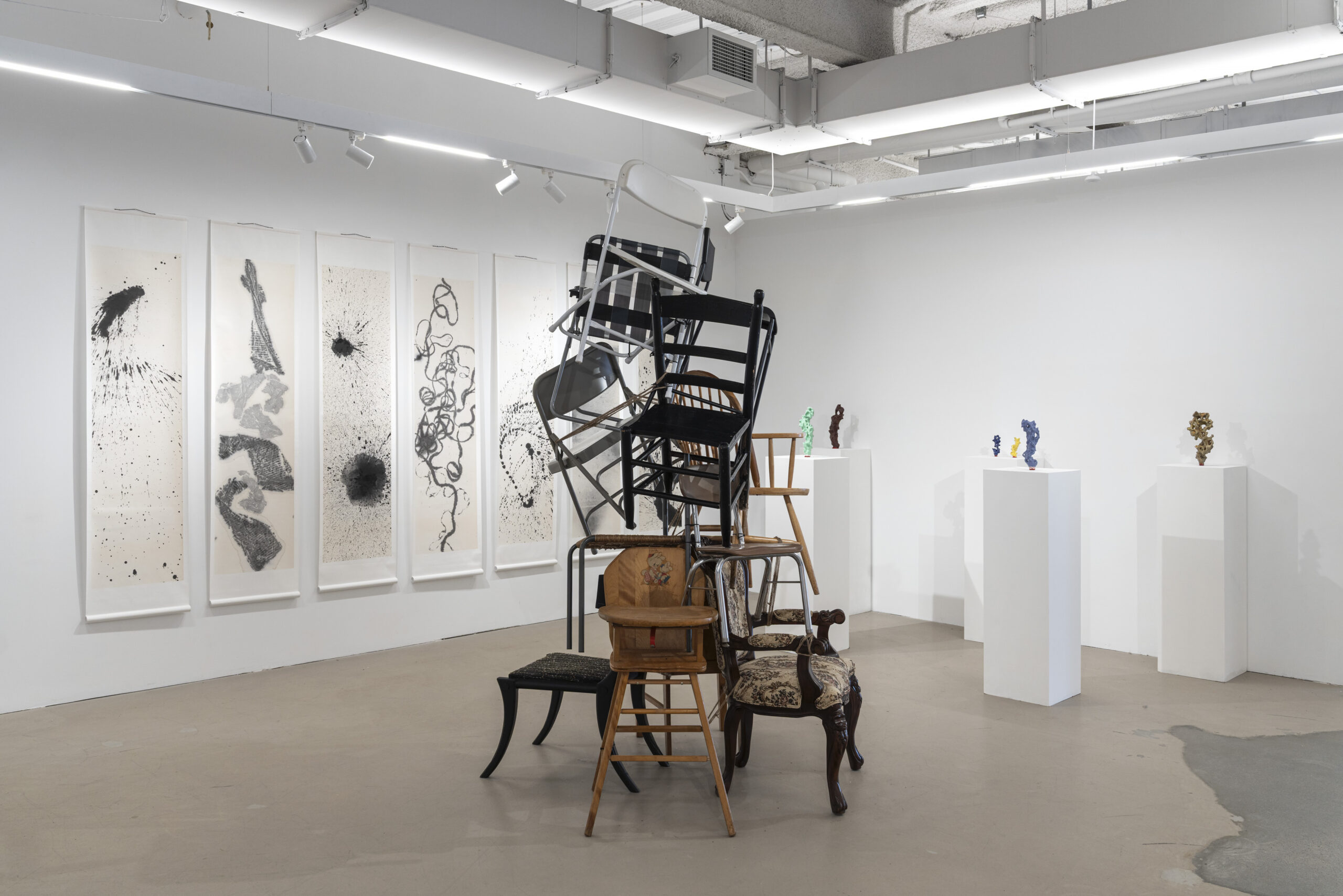 An art gallery interior featuring a central installation of multiple stacked chairs, including wooden and metal designs. In the background, five large framed panels exhibit abstract black ink splatter artwork. To the right, several small colorful sculptures are displayed on white pedestals against a plain wall. The space has bright lighting, highlighting both the artwork and the installations.