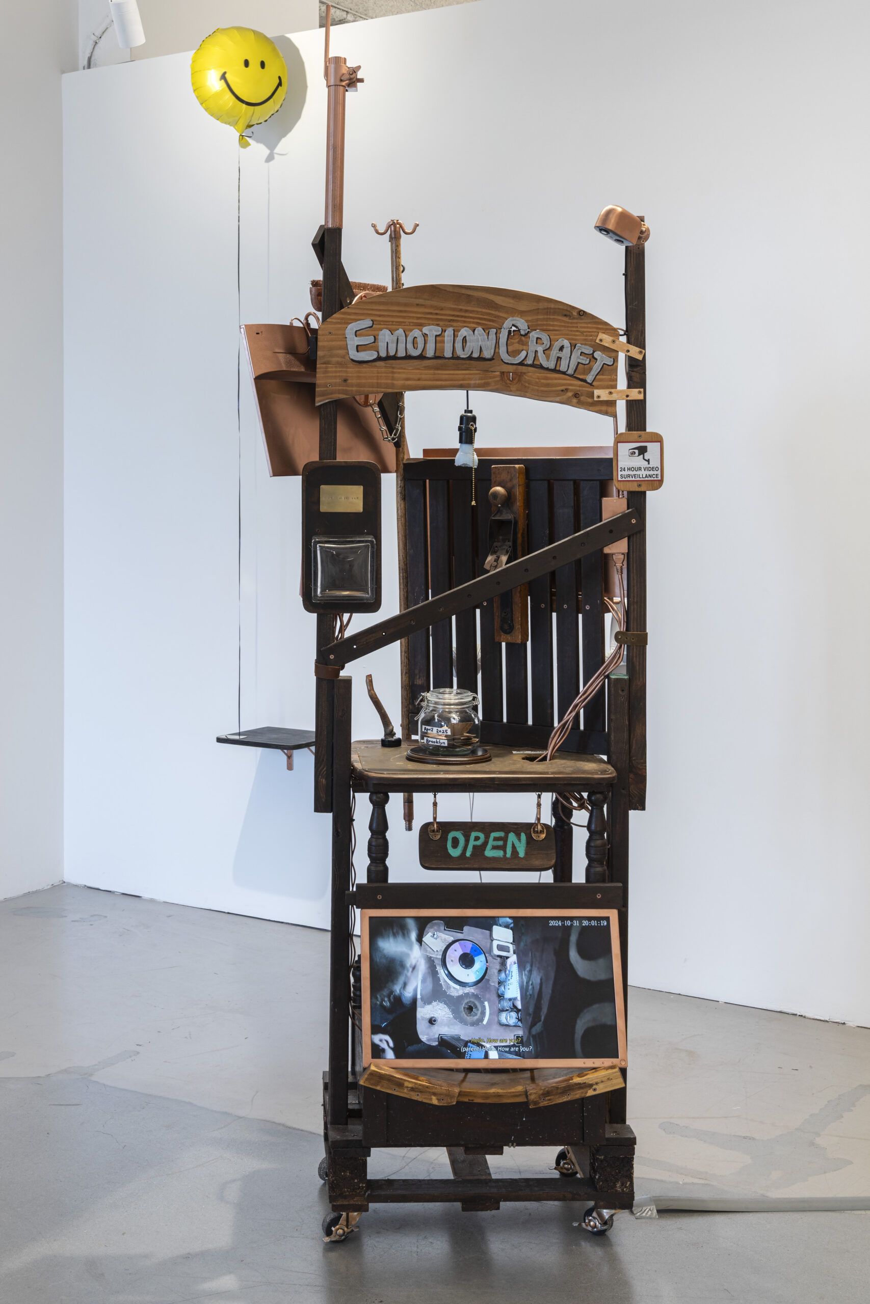 A quirky, artistic installation titled "Emotion Craft" stands in a minimalist gallery space. It features various elements, including a wooden sign above a collection of objects like an old-fashioned scale, light fixture, and glass jar. A large screen at the bottom displays an abstract close-up image. A bright yellow balloon with a smiley face floats above the structure, contributing to the whimsical theme. The overall design incorporates wood, metal, and contrasting textures.