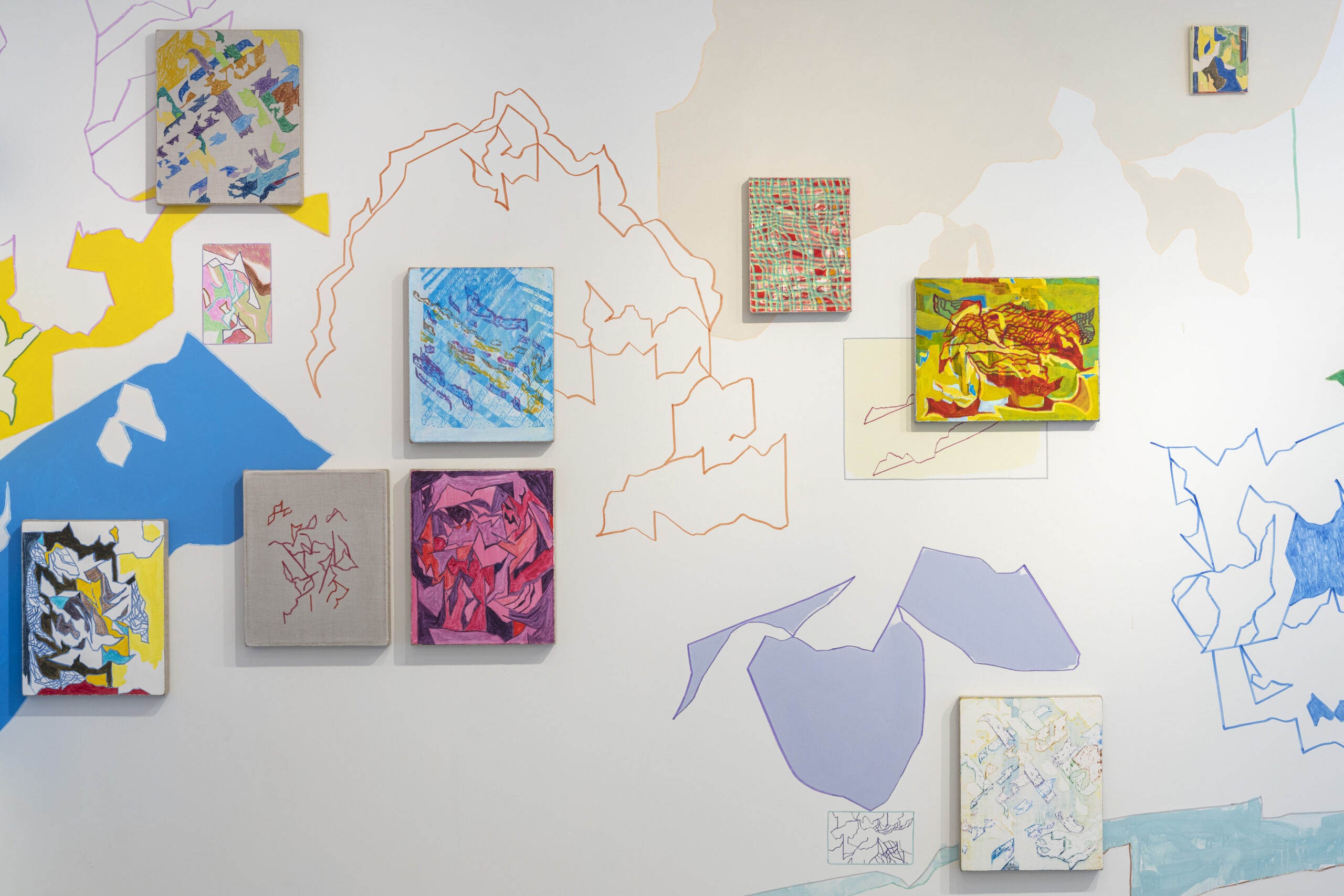 An array of colorful abstract paintings is displayed on a light wall, surrounded by vibrant painted shapes in blue, yellow, pink, and orange. The artworks vary in size and style, featuring bold lines and intricate patterns. Some paintings are bordered by softly colored outlines that match the wall's painted shapes. The overall aesthetic is playful and modern, creating a dynamic visual experience.