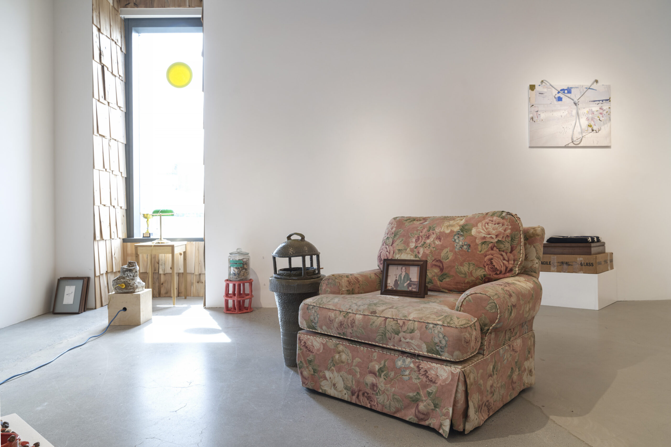 A brightly lit gallery with a floral-patterned armchair with a framed picture on its seat. Nearby, a decorative lantern is placed on a textured container, and a small table with a green lamp and a stone sculpture sits beside a window with wooden paneling. There’s also a low, light-colored table with a stack of boxes behind the chair, and a piece of artwork displayed on the wall. Sunlight streams into the room.
