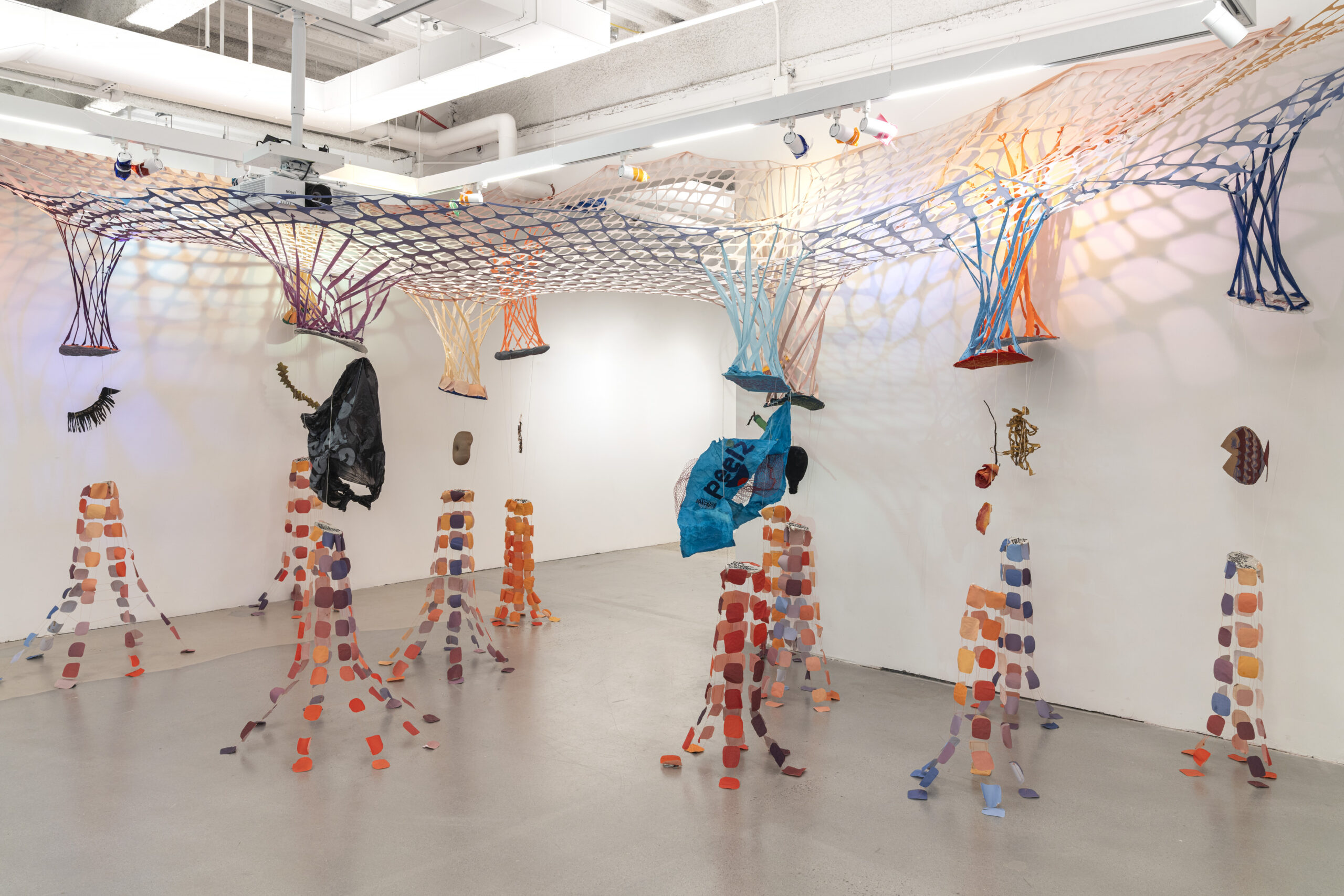 An art installation featuring colorful nets and sculptures suspended from the ceiling. The nets are in shades of blue, orange, and purple, casting shadows on the white walls. Below, there are vertical colorful strips resembling figures, with various objects like a black garment and painted shapes scattered throughout the space. The floor is light grey, and the overall atmosphere is vibrant and playful.