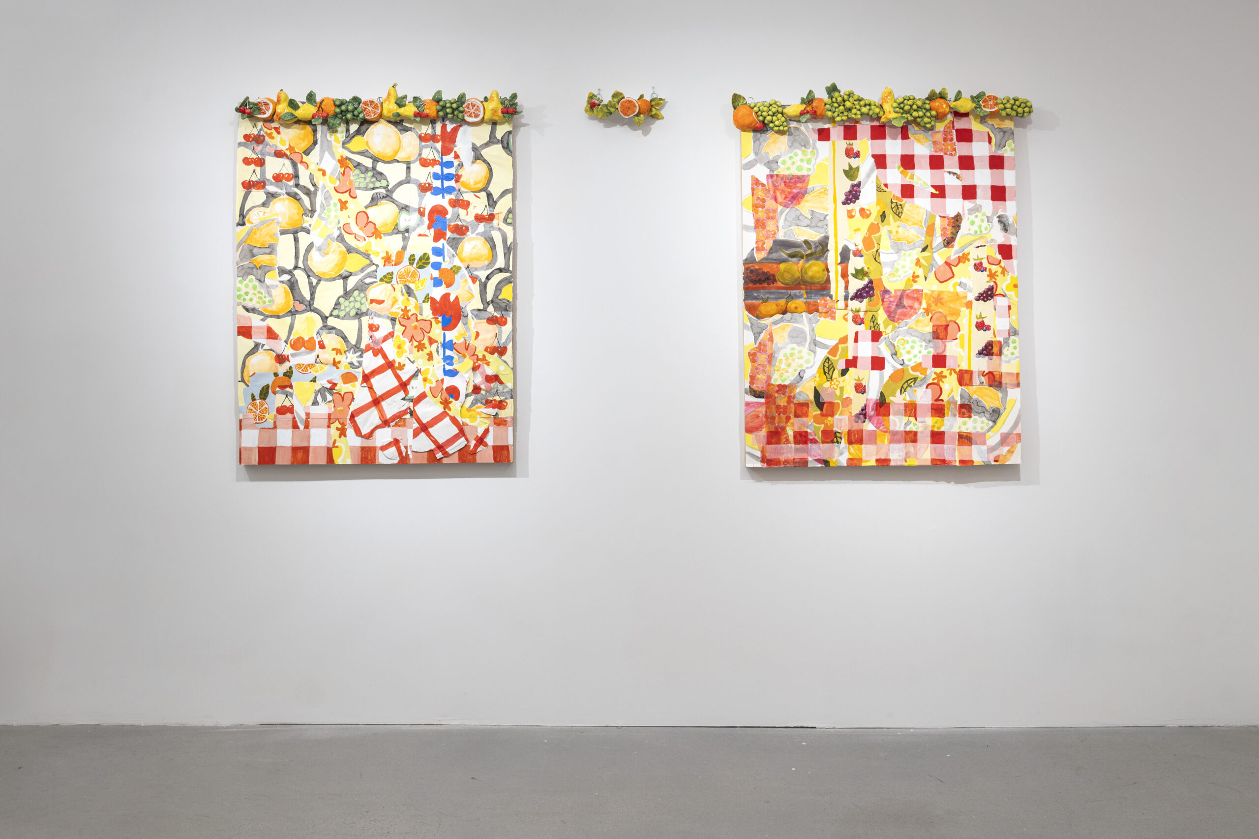 Two vibrant paintings hang on a white wall, featuring colorful fruits and floral patterns. The left painting has a red and white checkered pattern and showcases yellow lemons, tropical fruits, and various designs. The right painting also features a checkered background, filled with bright reds, yellows, and greens, depicting assorted fruits, vegetables, and abstract shapes. Both paintings are adorned with a decorative border of colorful faux fruits along the top.