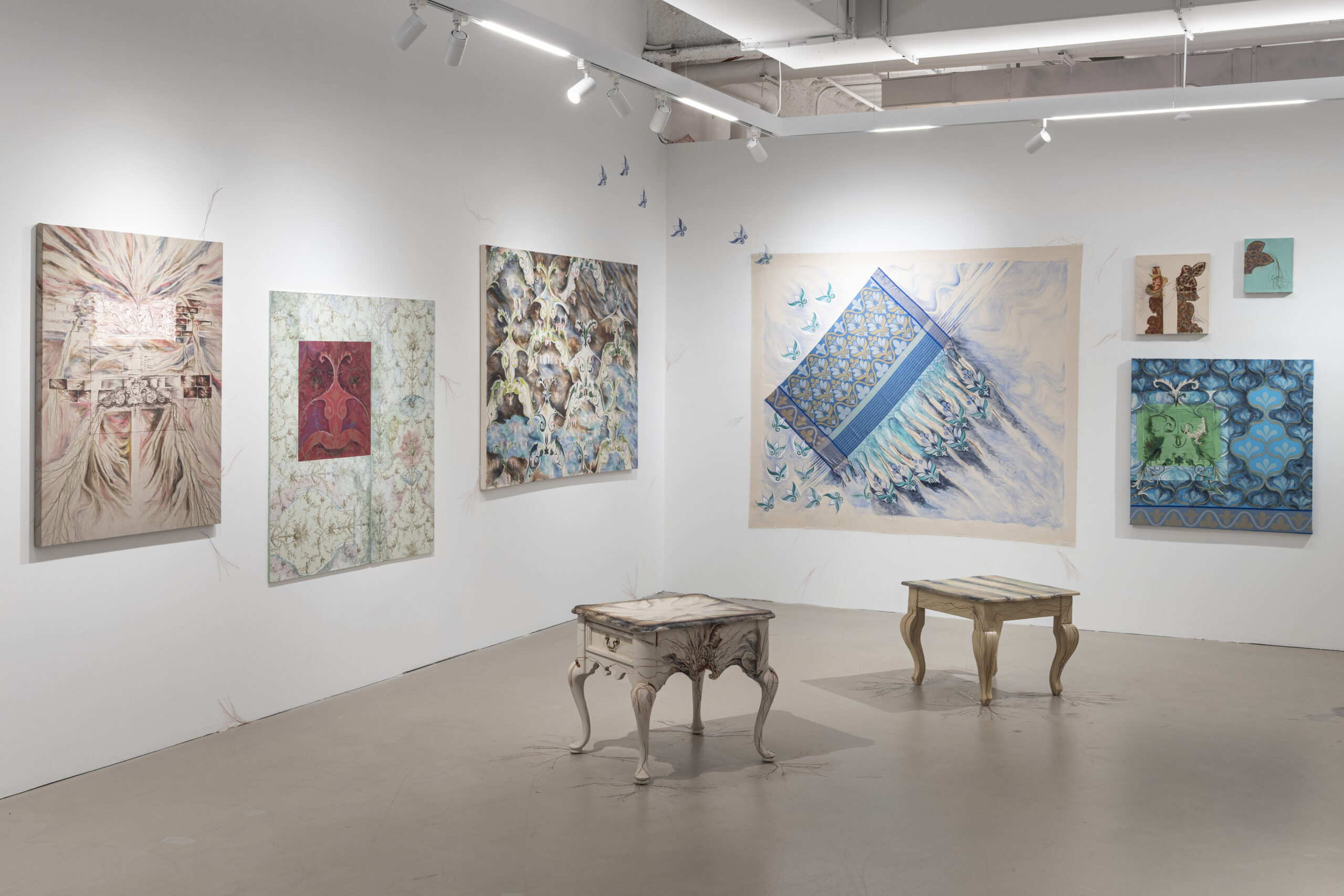 An art gallery interior featuring a two short, squat white tables in the foreground. On the left wall, there are four abstract pieces: a beige one, followed by a dark blue, then two red pieces in the format of raised shapes. To the right, there are two framed artworks, one blank and one with a blue design. The floor is a plain gray concrete surface.