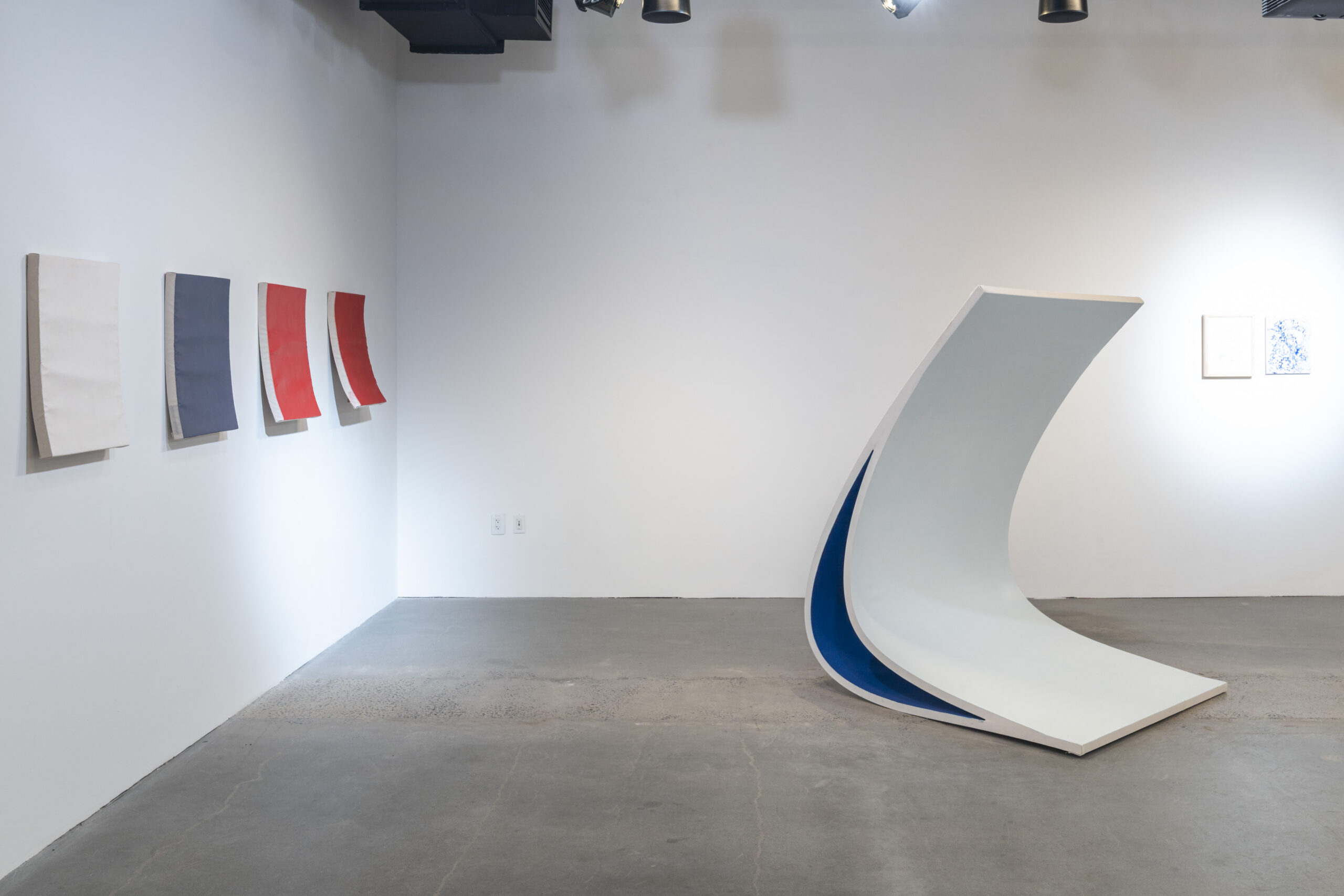 An art gallery displays a minimalist installation featuring four curvilinear wall pieces in shades of white, blue, red, and gray. In the foreground, a large, abstract white sculpture bends to reveal a blue interior. The polished concrete floor reflects ambient light from above, enhancing the contemporary aesthetic of the space. Two framed artworks are visible on the wall to the right.