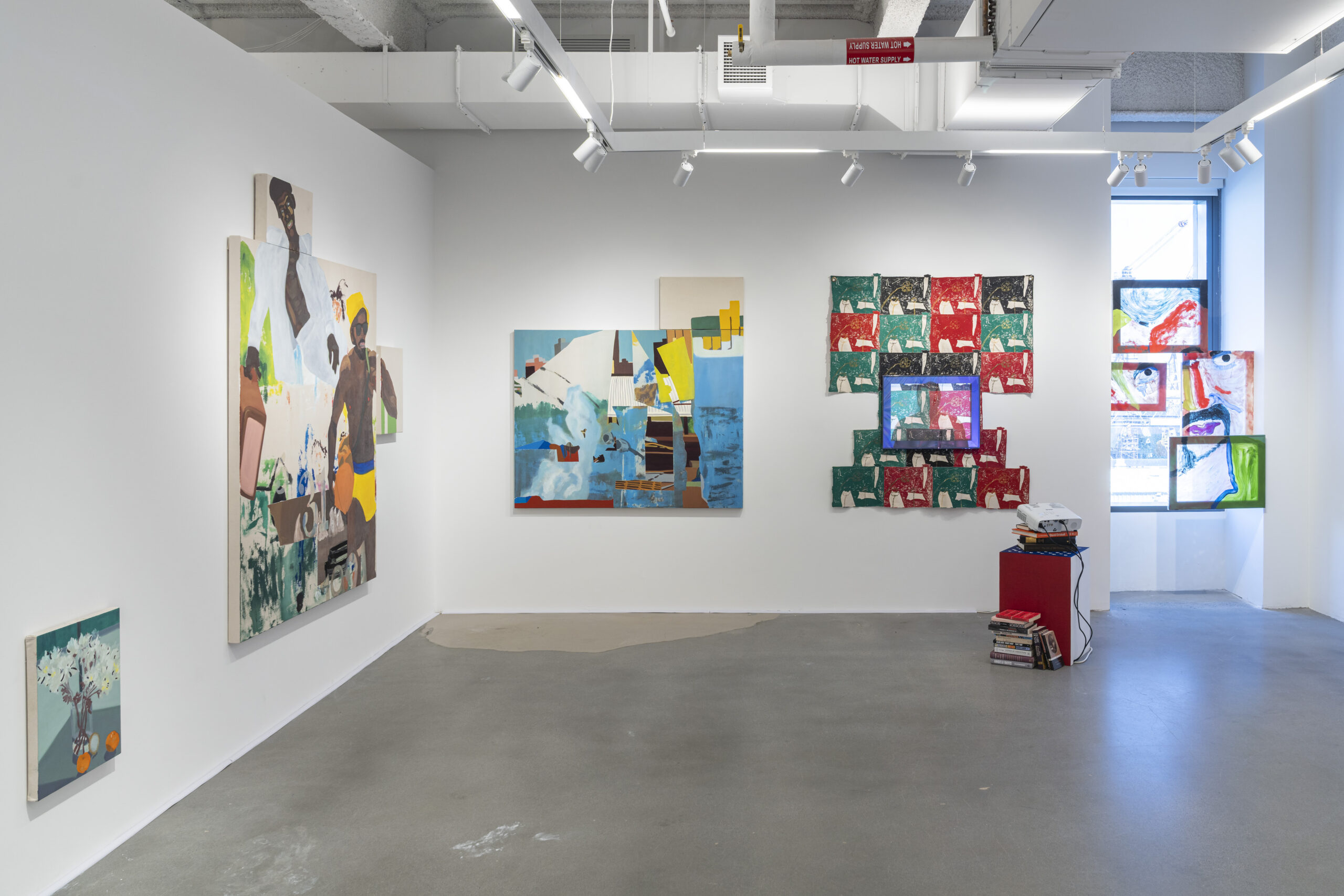 An art gallery interior features several artworks displayed on white walls. To the left, a painting depicts a figure with a yellow swimsuit amidst abstract elements. A central large artwork combines bright colors and shapes. To the right, a series of patterned pieces in red, green, and black creates a layered visual effect, backlit by a window. A small red pedestal holds a stack of books and a projector, and a window allows natural light to illuminate the space.