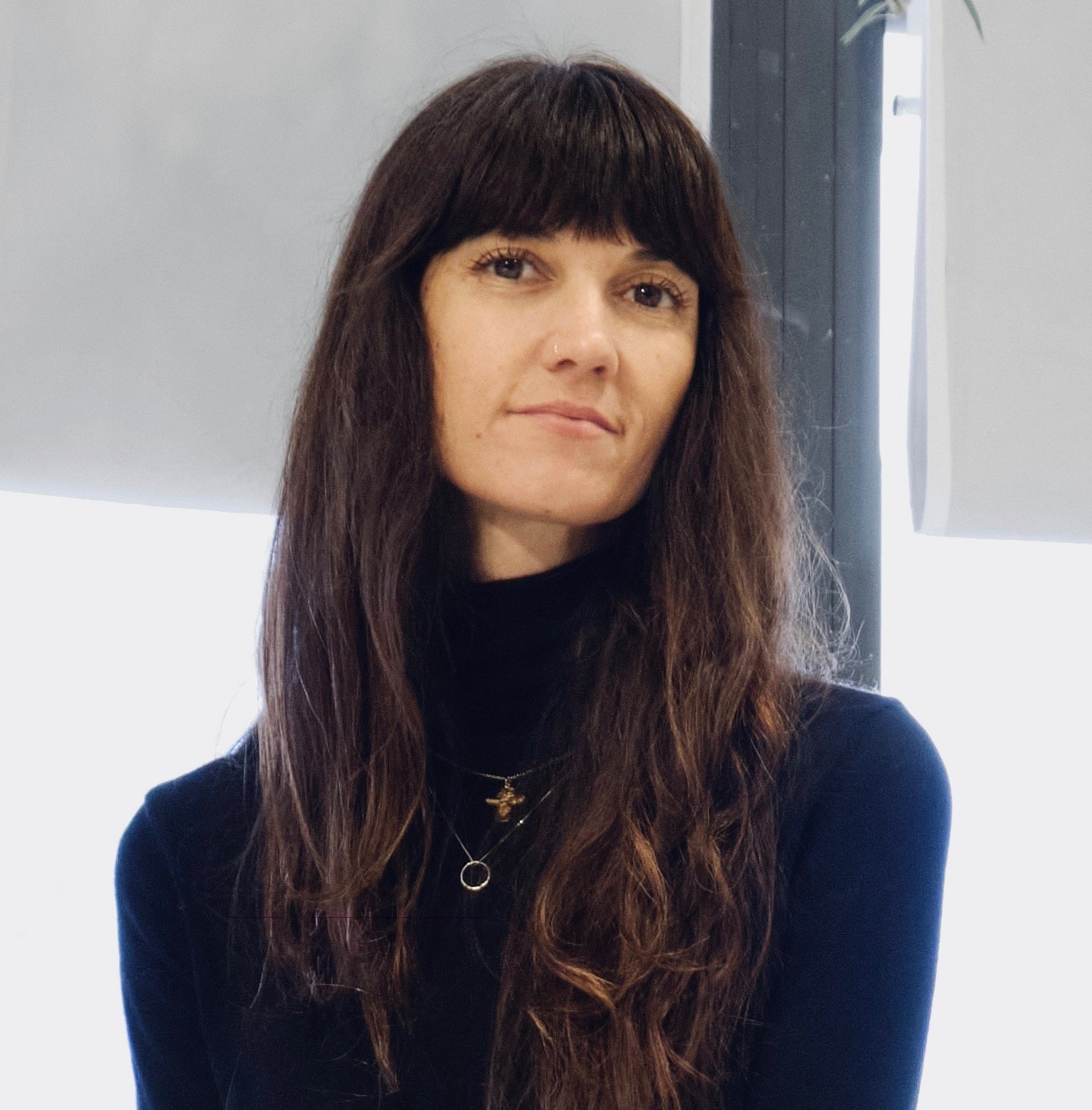 Portrait of Langdon Graves long, dark hair and bangs with minimal natural light. She is wearing a black turtleneck and have a serene expression. Small gold necklaces are visible around their neck. The background features blurred elements.