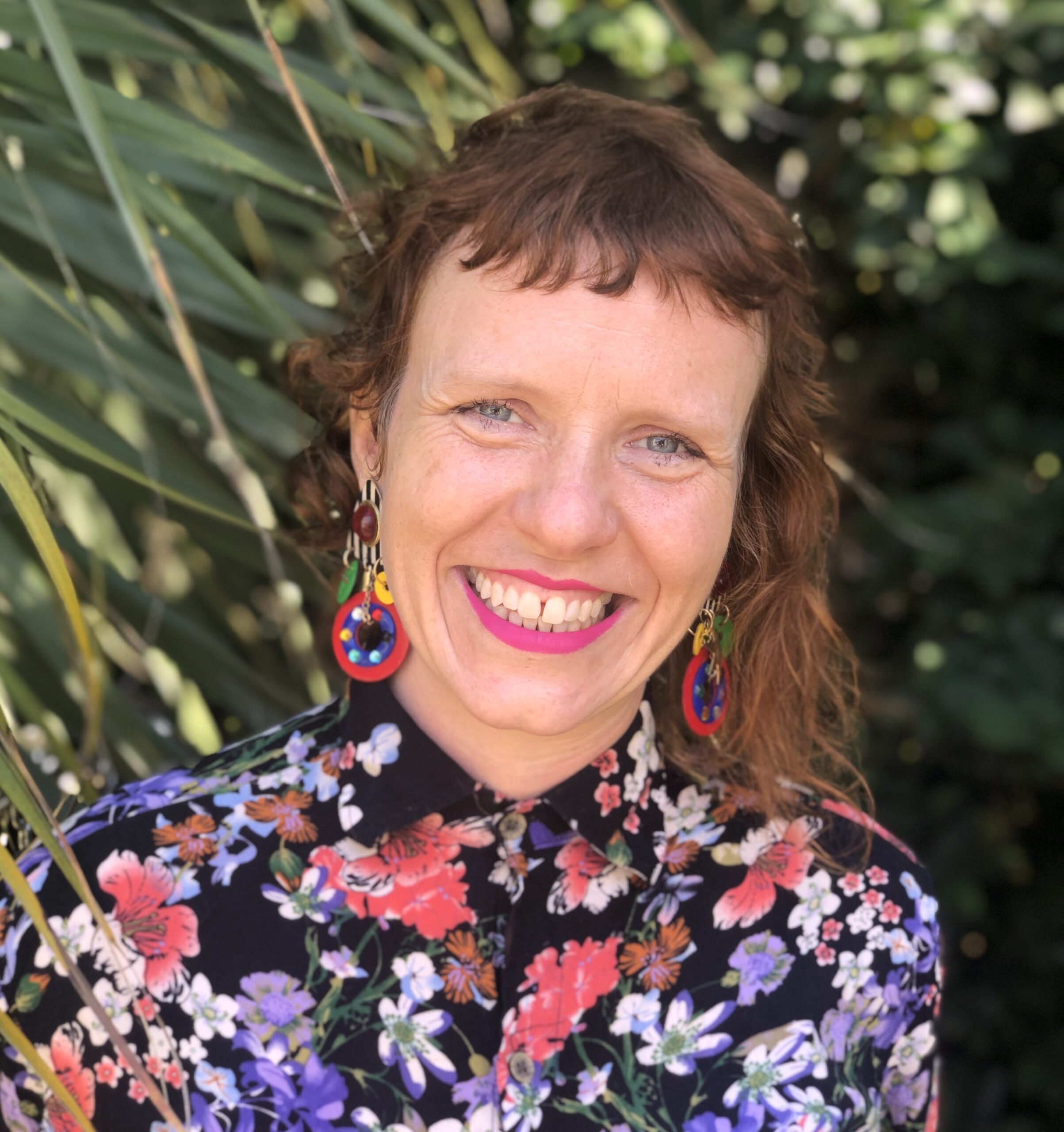 A person with long, dark hair is wearing a black turtleneck and is standing indoors. They are smiling and looking directly at the camera. They are wearing a brightly colored floral shirt, bright earrings, and standing in front of greenery and foliage