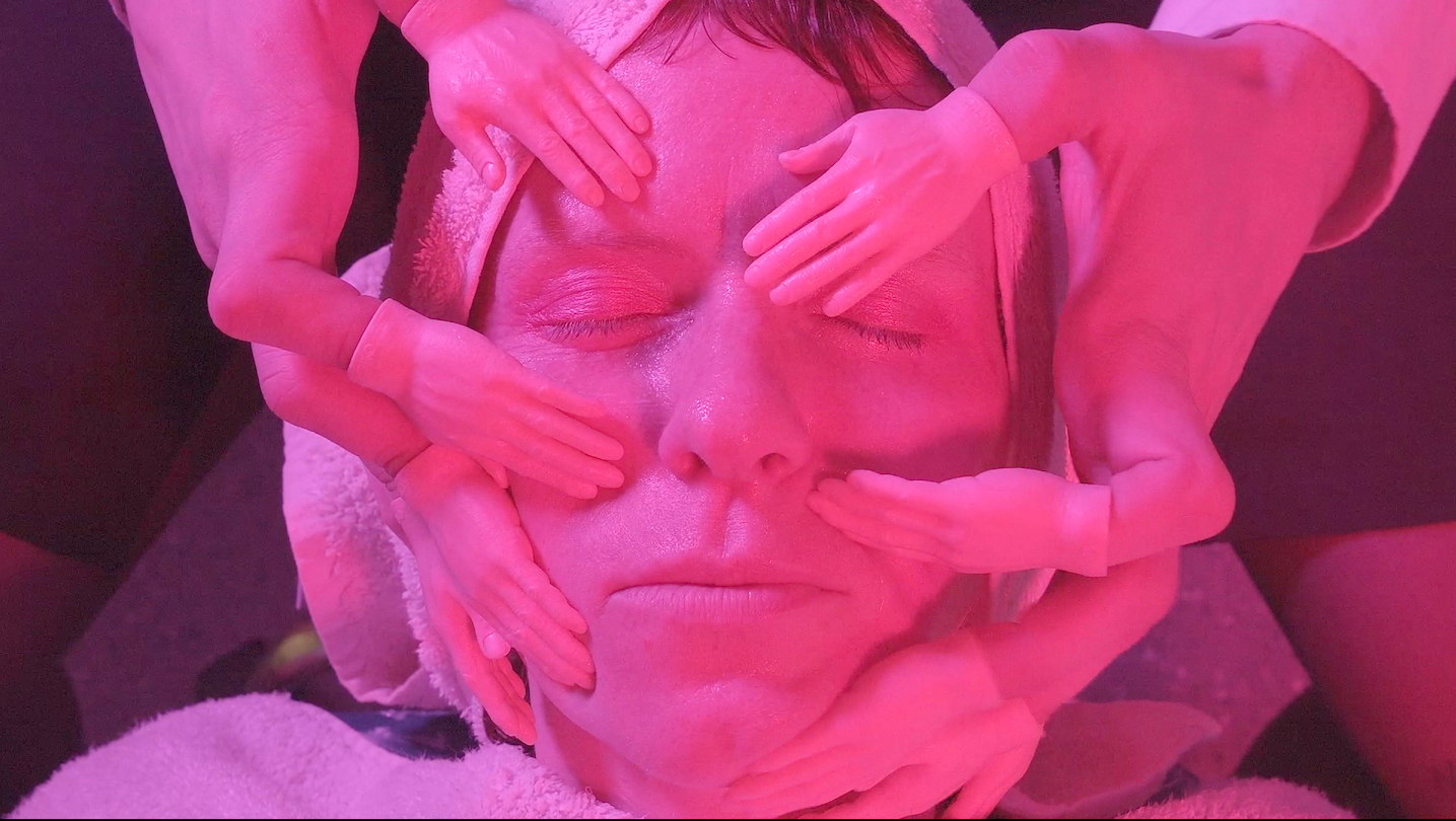 A close-up of a person's face, eyes closed, surrounded by several small hands made from a doll or figurine, which appear to be gently touching their face. The scene is illuminated with pink lighting, creating a surreal atmosphere. The person has a towel wrapped around their head, and a white towel is visible in the background.