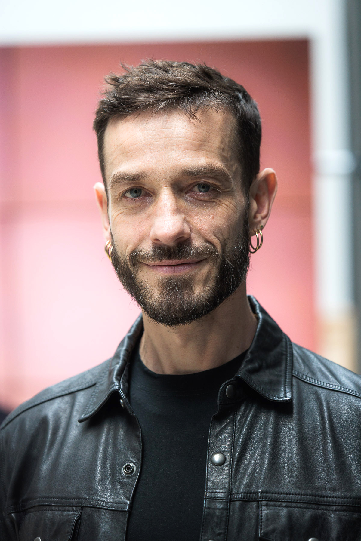 A man with short, dark hair and a well-groomed beard smiles at the camera. He is wearing a black leather jacket over a black t-shirt. His left ear has a hoop earring and he has a confident expression. The background features soft, blurred pink tones.