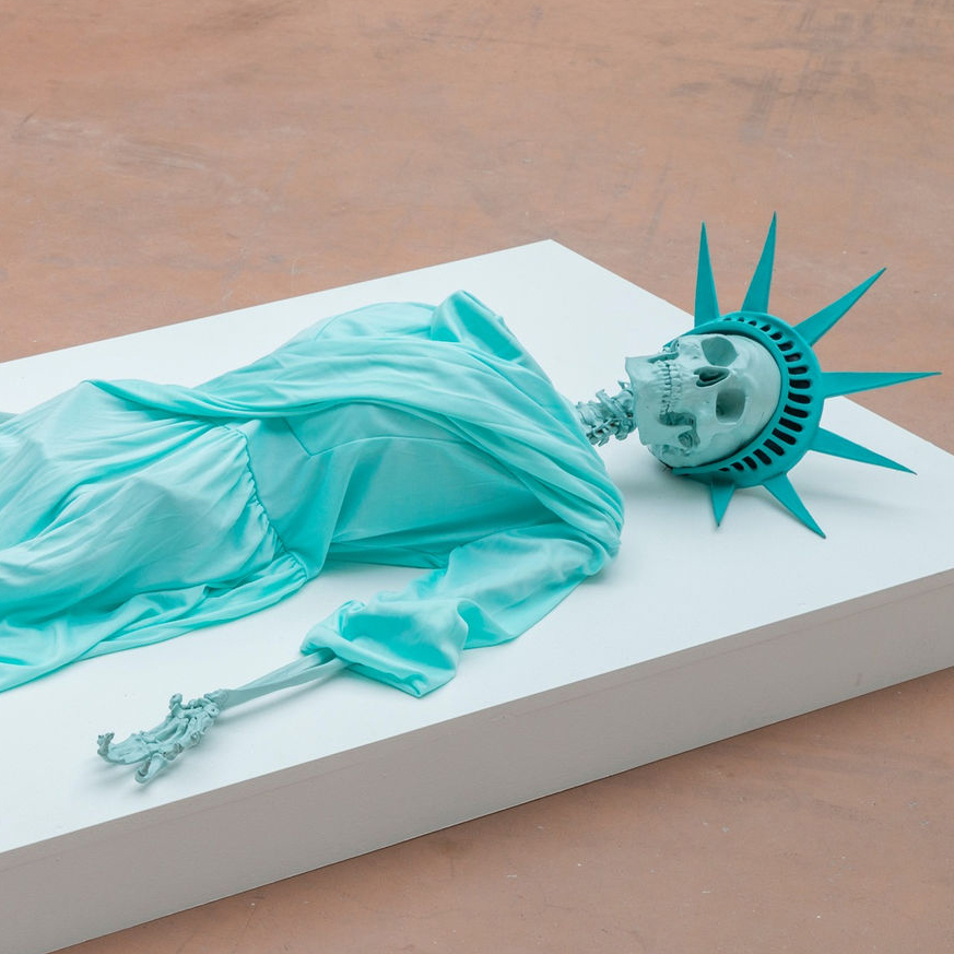 Skeleton painted green wearing a Statue of Liberty Costume laying horizontally on a low white platform on a light brown floor.