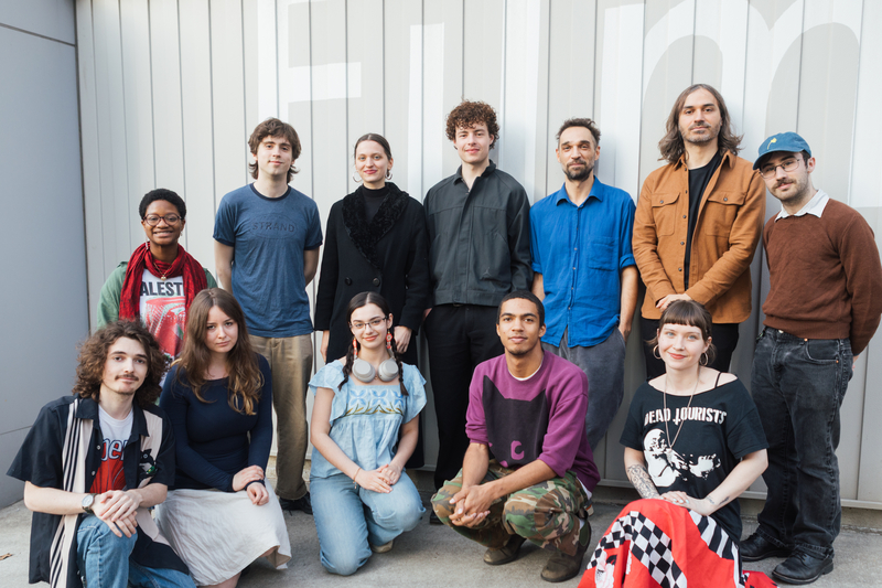 A group of fourteen individuals poses for a photo outdoors in front of a wall with the word "Film." They are arranged in two rows; the back row features seven people standing, while the front row has seven people seated. The group exhibits a variety of clothing styles and expressions, suggesting a casual and friendly atmosphere.