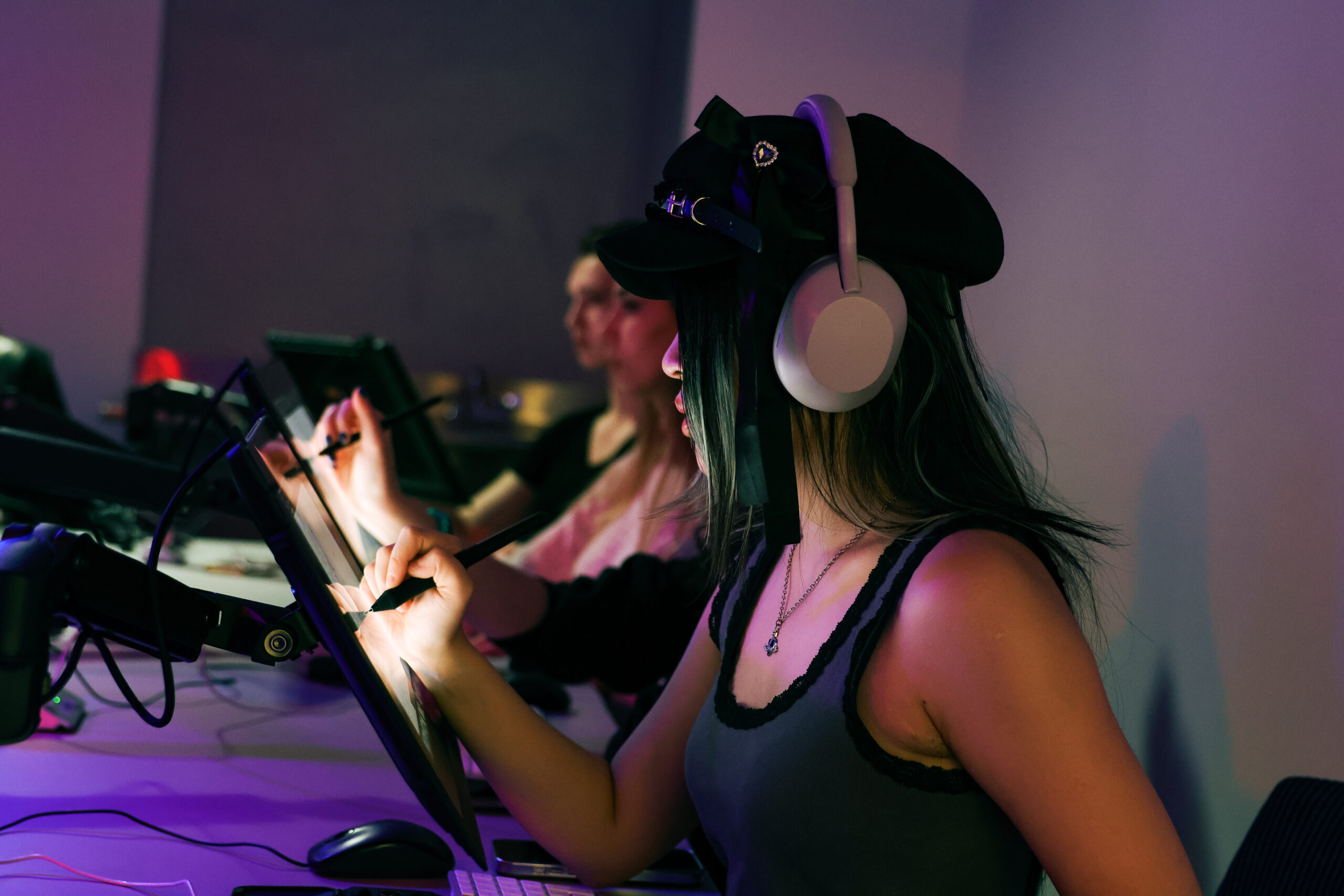 Students work at digital pen display workstations during a drawing class. In the foreground, a student wearing over-ear headphones concentrates on sketching with a stylus while another student draws at a neighboring workstation in the softly lit studio.