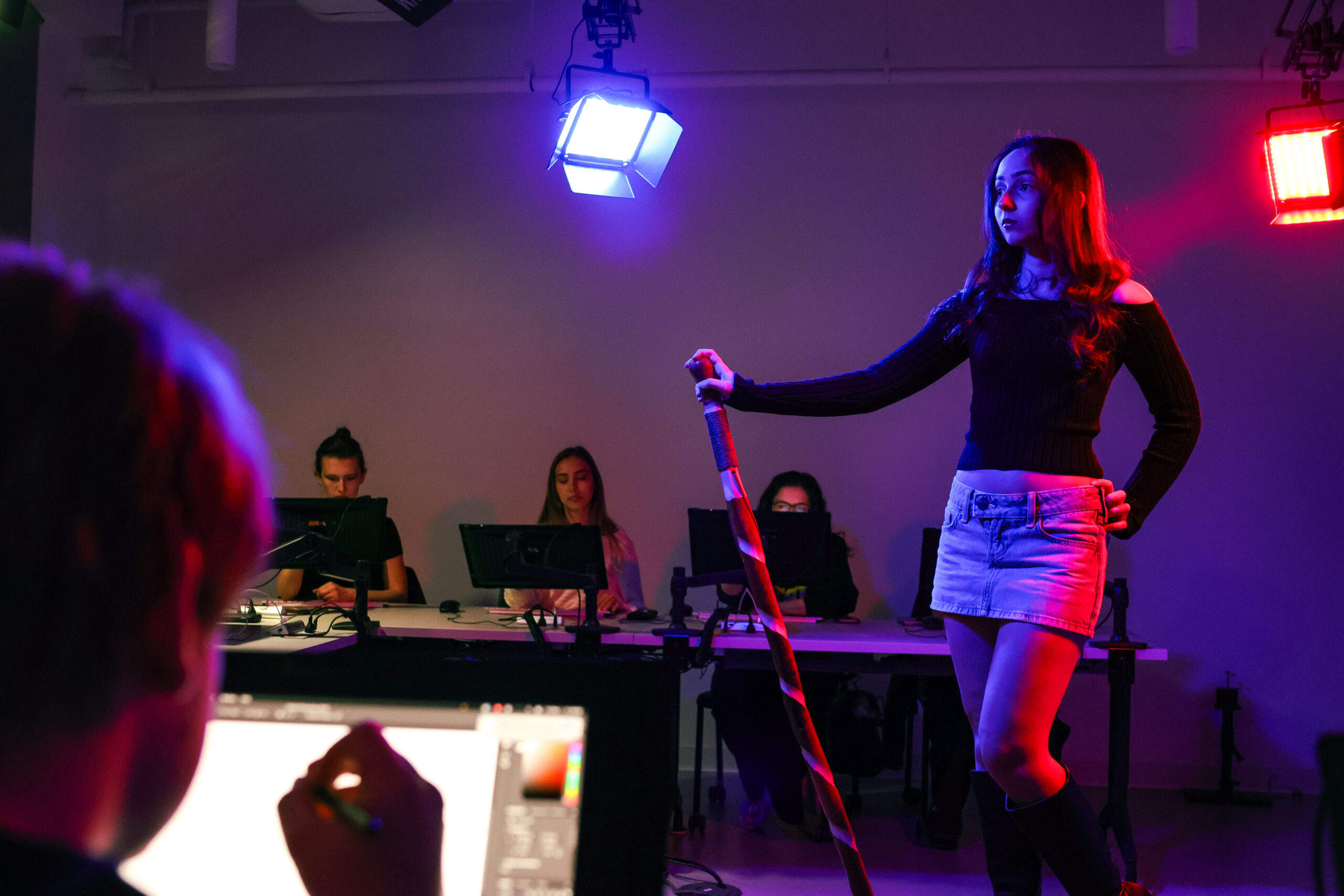 A student poses under blue and red studio lights while classmates observe and draw from reference at digital workstations. The model stands holding a staff as students practice figure drawing using pen displays in a digital art studio.