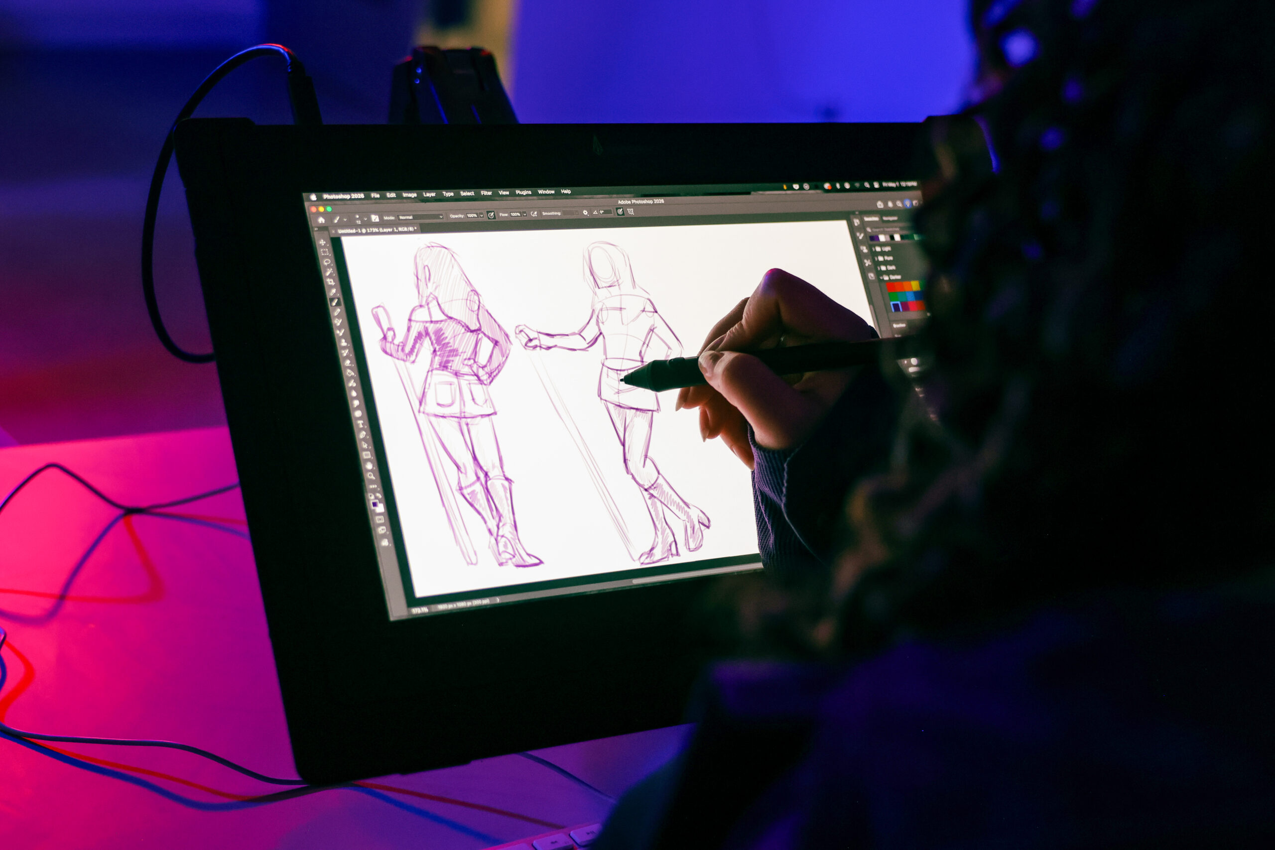 A student uses a digital pen display to sketch two character pose studies in Adobe Photoshop. Purple line drawings of a figure holding a staff are visible on the screen, illuminated by colorful studio lighting in a digital drawing classroom.