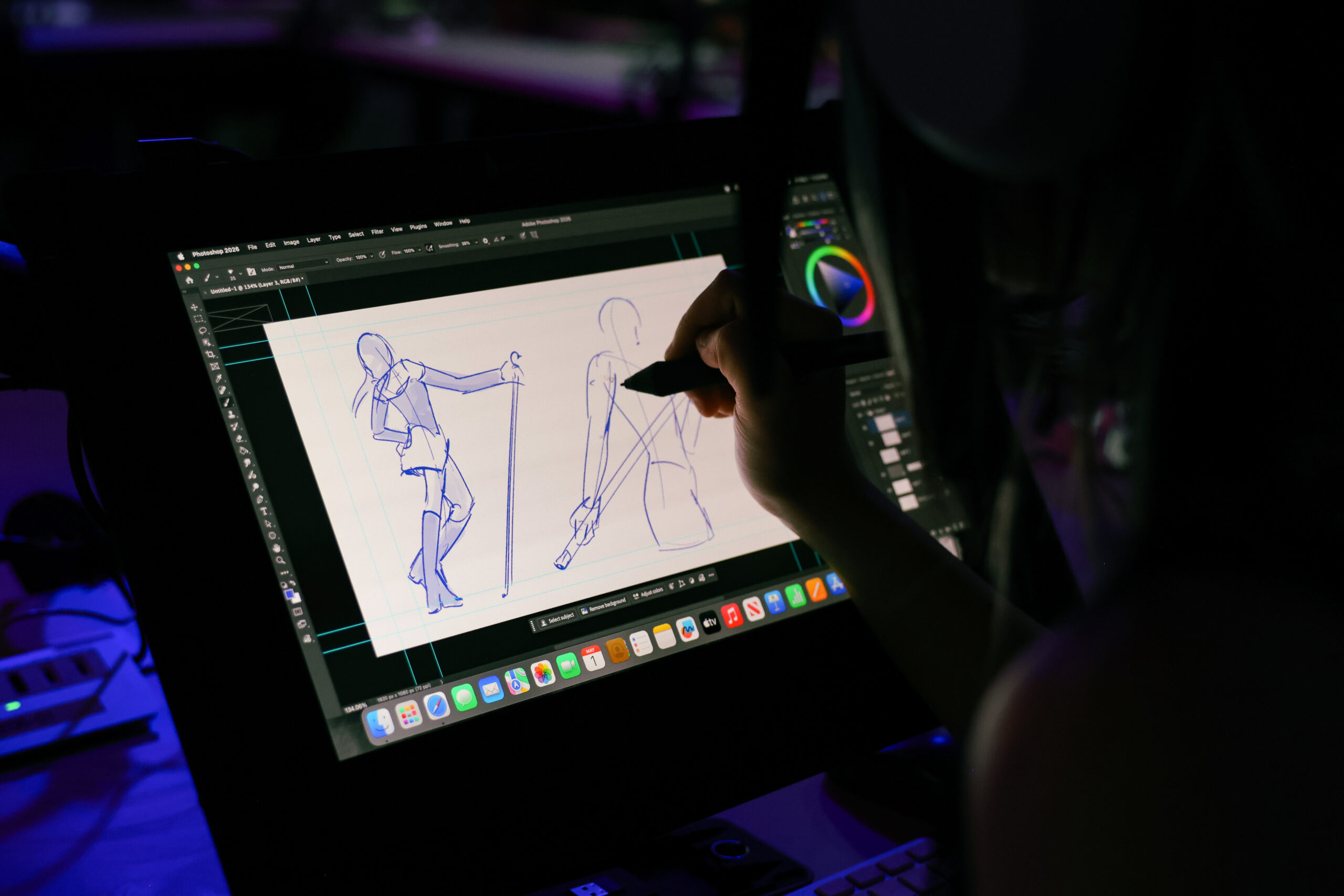 A close-up shows a student drawing character gesture studies with a stylus on a digital pen display in Adobe Photoshop. Blue sketch lines depict a standing figure holding a staff, with the workstation illuminated by low ambient lighting.