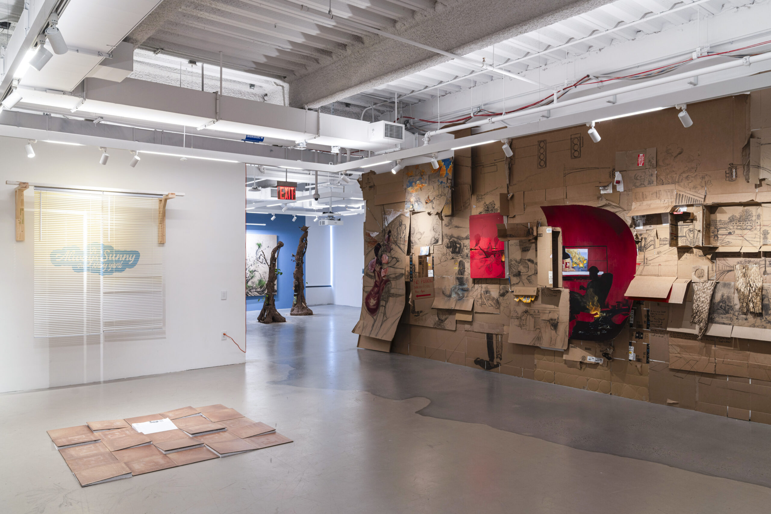 An art gallery interior featuring two main installations. To the left, there is a wall with sheer blinds and a sign that reads "Sunny." On the right, a large cardboard installation displays drawings and a bright red panel with a scene depicting a figure in front of a television. Scattered across the floor are several flat tiles arranged in a patchwork layout. In the background, a blue wall showcases other artworks, including two tree-like sculptures.