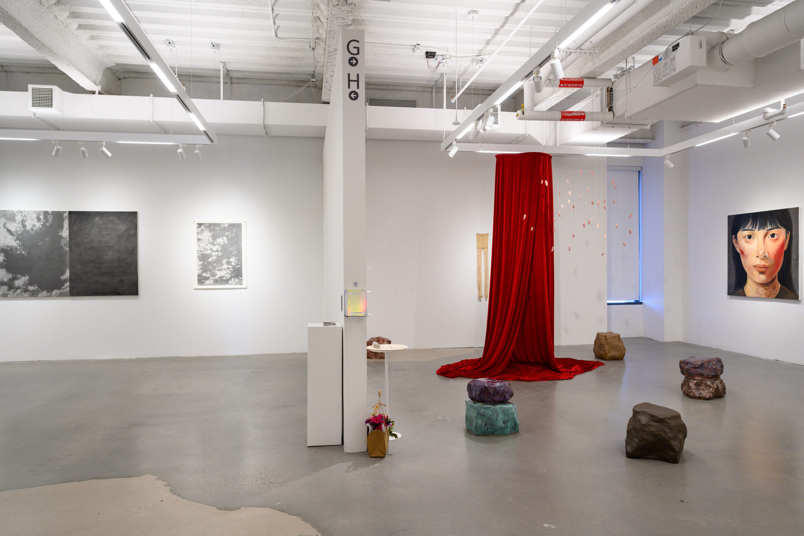 A contemporary art gallery showcasing a large red curtain at the center, with hanging decorative elements above. On the walls are various artworks, including two black-and-white drawings and a portrait of a woman. Scattered around the floor are multi-colored stones and a small pedestal with a reflective object. The lighting is bright, highlighting the artwork and features of the gallery space.