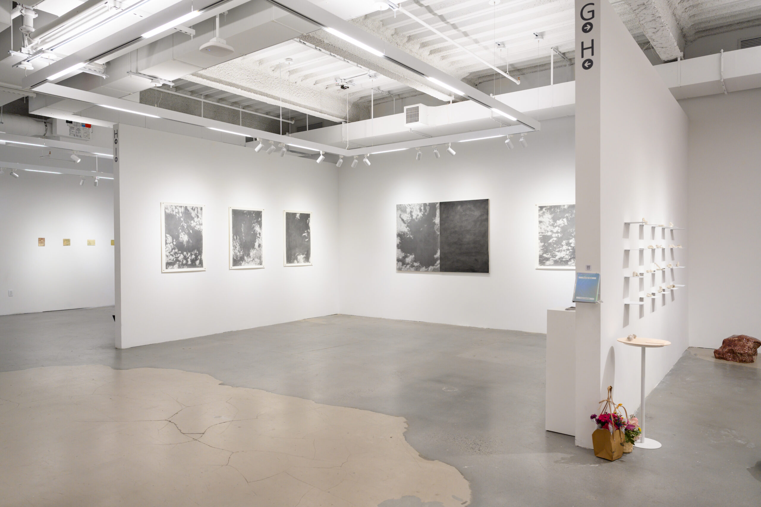 A contemporary art gallery with white walls and a polished concrete floor. Several artworks are displayed, including black-and-white drawings on the left wall. On the right side, there are shelves featuring small sculptures or objects. A small table with flowers and a bag is positioned nearby, and a digital screen is on a pedestal. Bright lighting is overhead, enhancing the artwork's visibility.