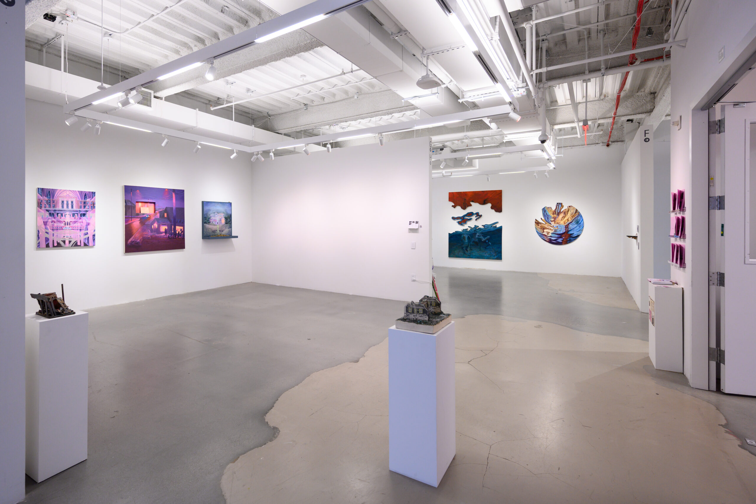 An art gallery interior featuring several artworks on bright white walls. On the left, two paintings with bold colors and abstract forms are displayed, with a small sculpture on a pedestal nearby. In the center, three more artworks are visible—one abstract piece in red tones, another in deep blues, and a circular painting with earthy colors. The floor is a mix of gray and white concrete, and there is a door leading to another area on the right.