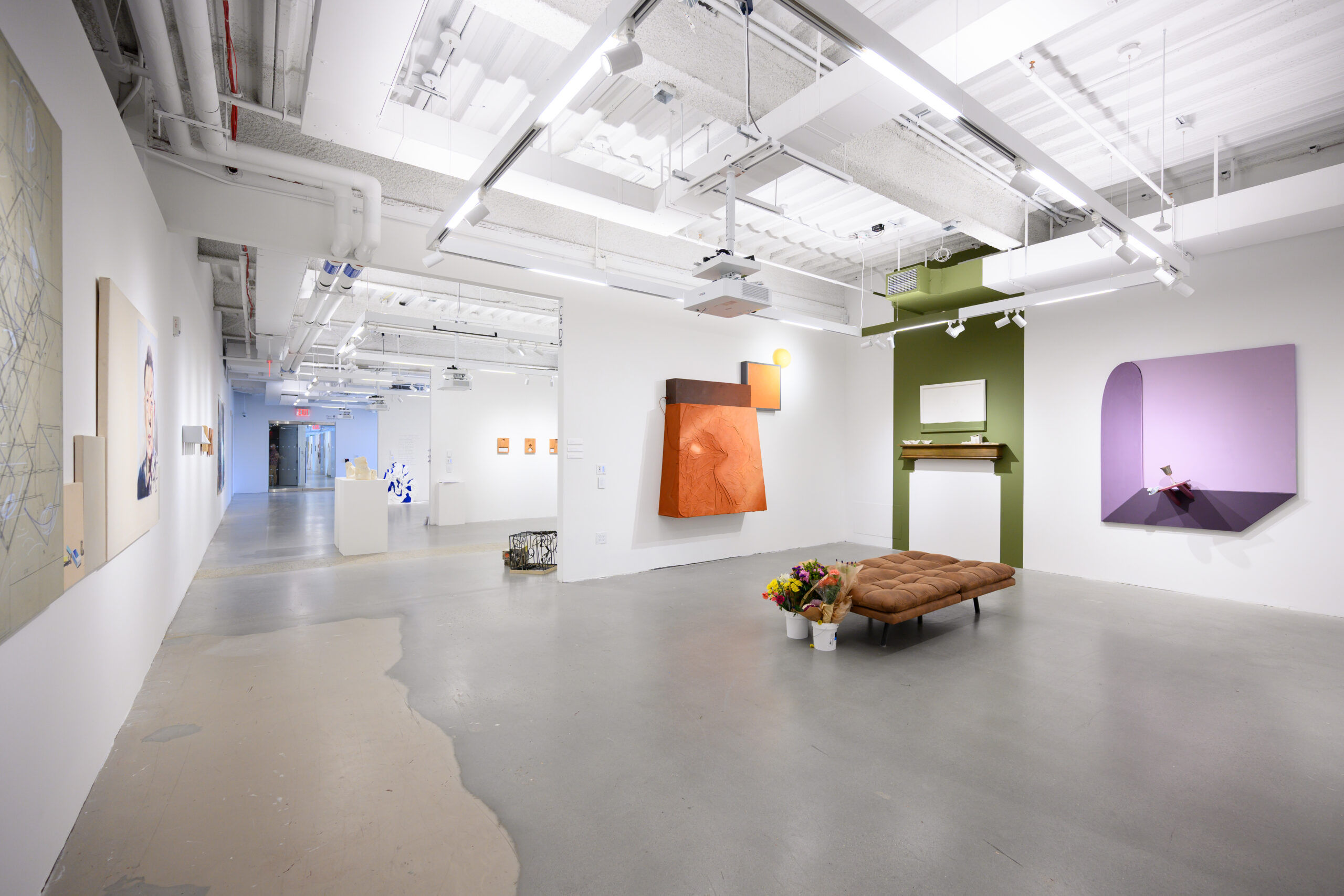 An art gallery interior features white walls displaying various artworks. A large, textured orange piece hangs on the wall, complemented by a purple painting nearby. A green area with a shelf and a mirror adds contrast. In the center, a brown tufted futon is surrounded by flower arrangements placed in white containers. The floor is a polished concrete surface with light reflections.