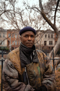 A man stands outdoors, wearing a dark hat and a patterned, oversized jacket. He has a serious expression and is positioned in front of a backdrop featuring bare trees and residential buildings. The scene is set in a park-like area, suggesting a cool, overcast day.