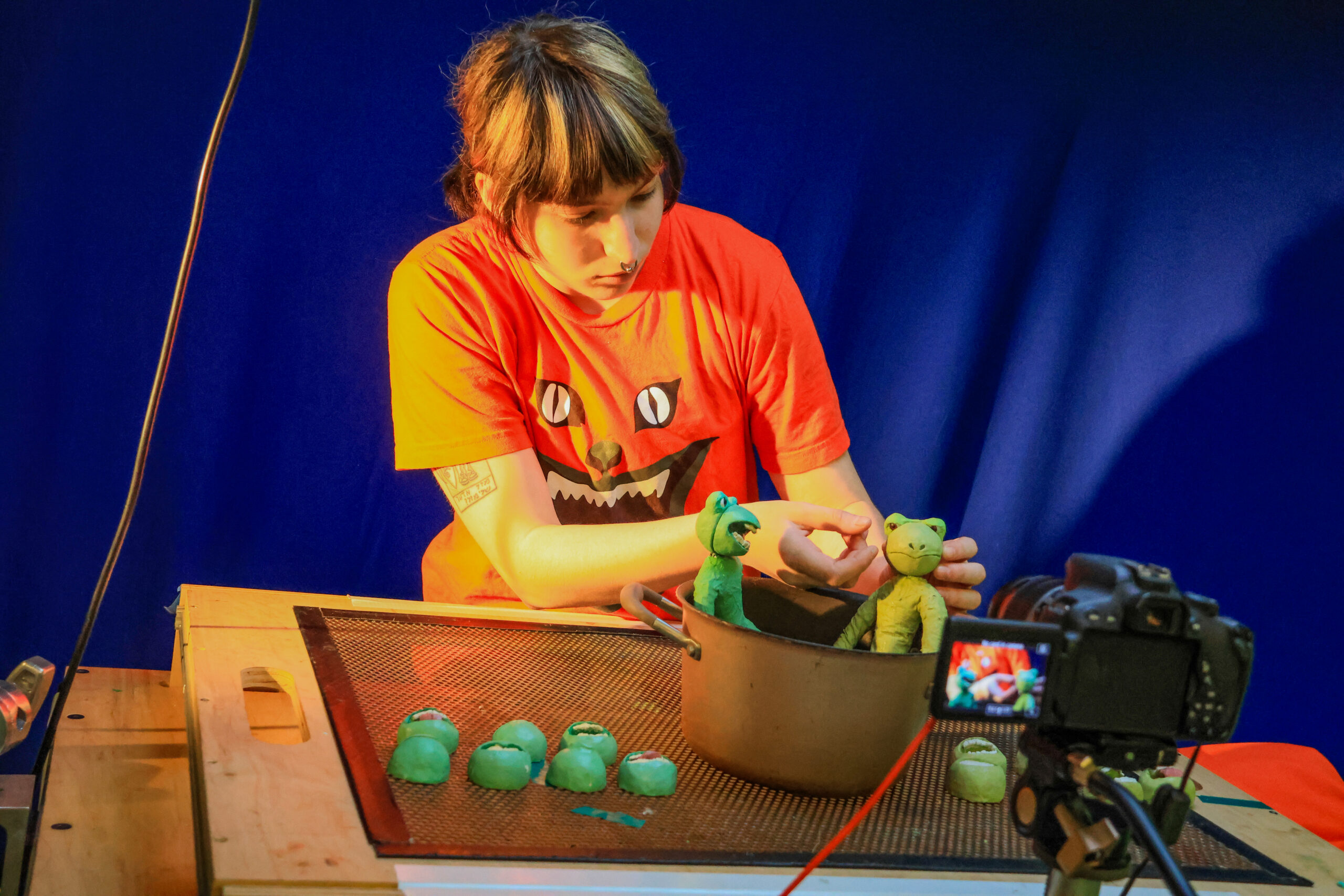 A student animates two green clay characters on a tabletop stop-motion set, carefully positioning one character inside a metal pot while a DSLR camera records the scene under colored studio lighting against a blue backdrop, with replacement clay parts arranged nearby.