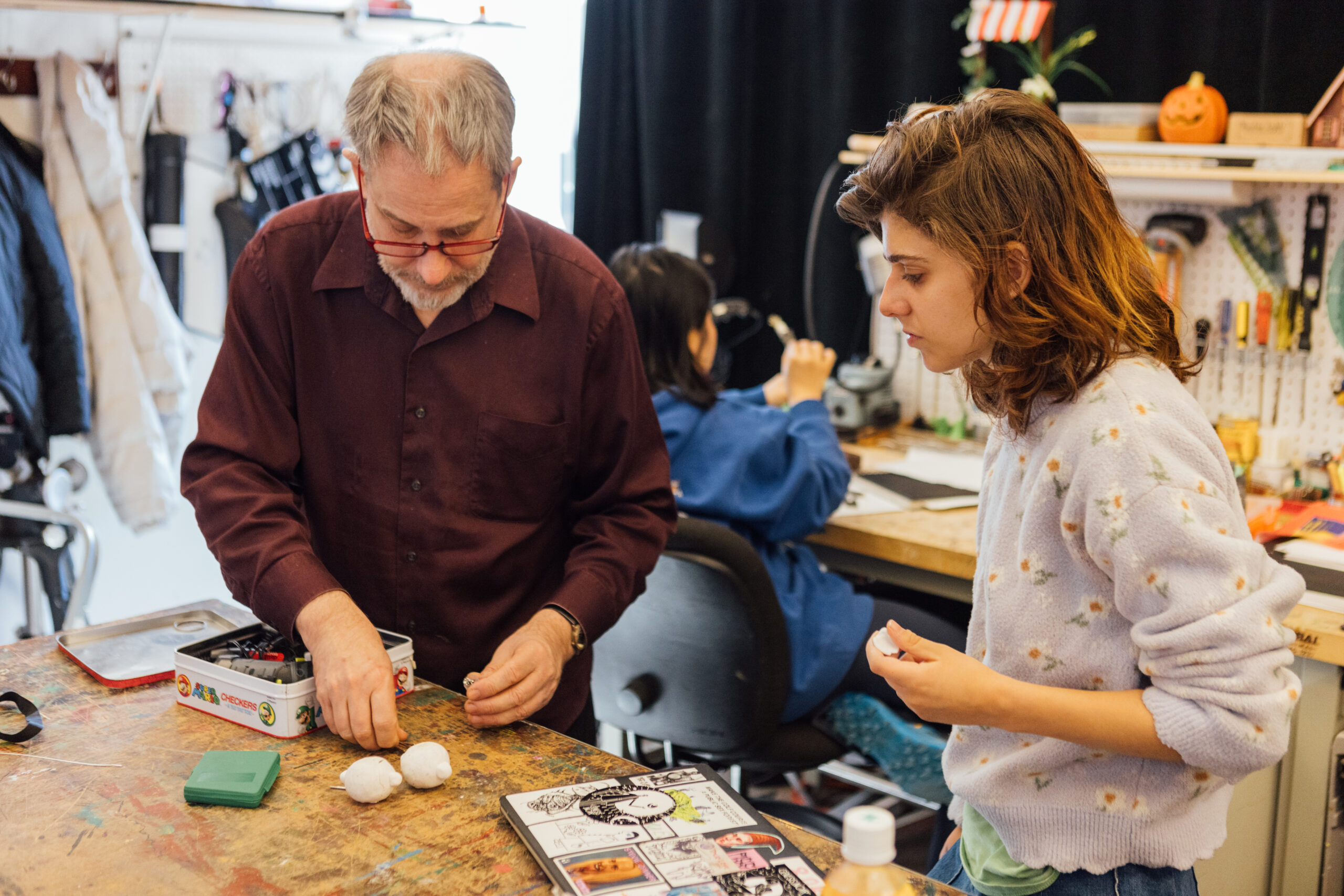 An instructor demonstrates stop-motion animation techniques with clay models at a workbench while a student observes closely, as another student works at a station in the background surrounded by tools, materials, and animation equipment in a studio classroom.