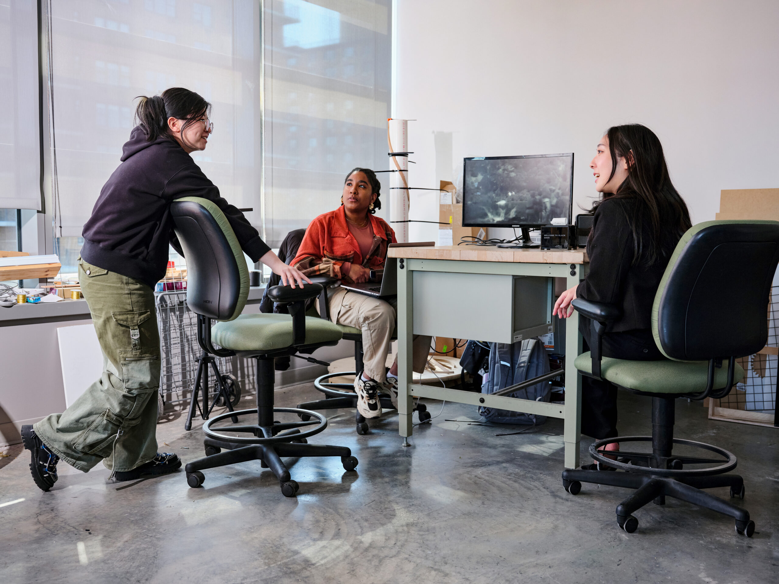 Three students converse around a worktable in a digital arts studio, with one student standing and leaning on a chair while two others sit beside a computer workstation, discussing a project in a bright workspace with large windows and studio materials.