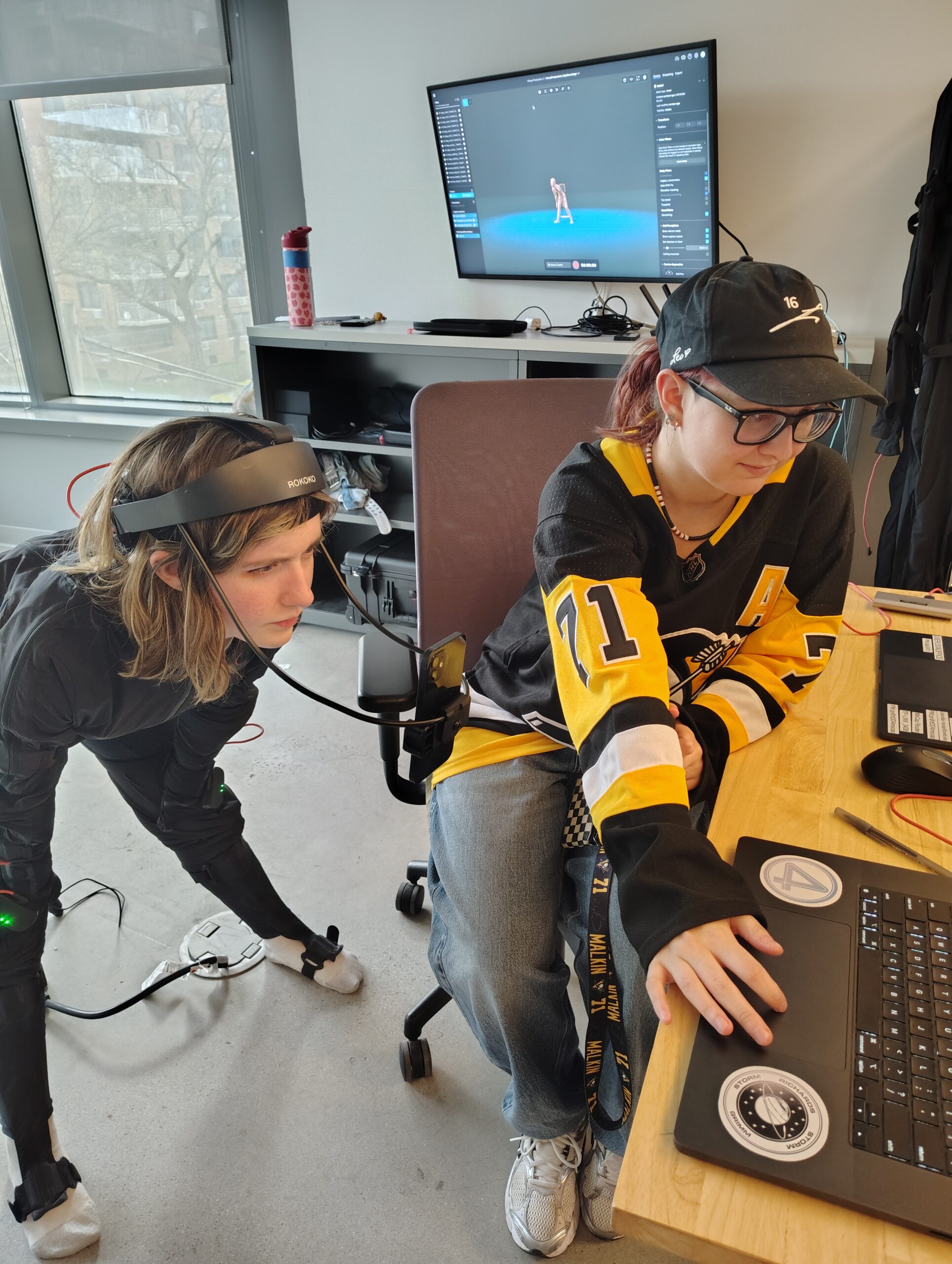 Two students work together on a motion capture project at a workstation. One student wears a motion capture suit and head-mounted tracking device while leaning toward the computer, as the other student operates the software on a laptop. A monitor in the background displays a 3D character generated from the captured motion data.