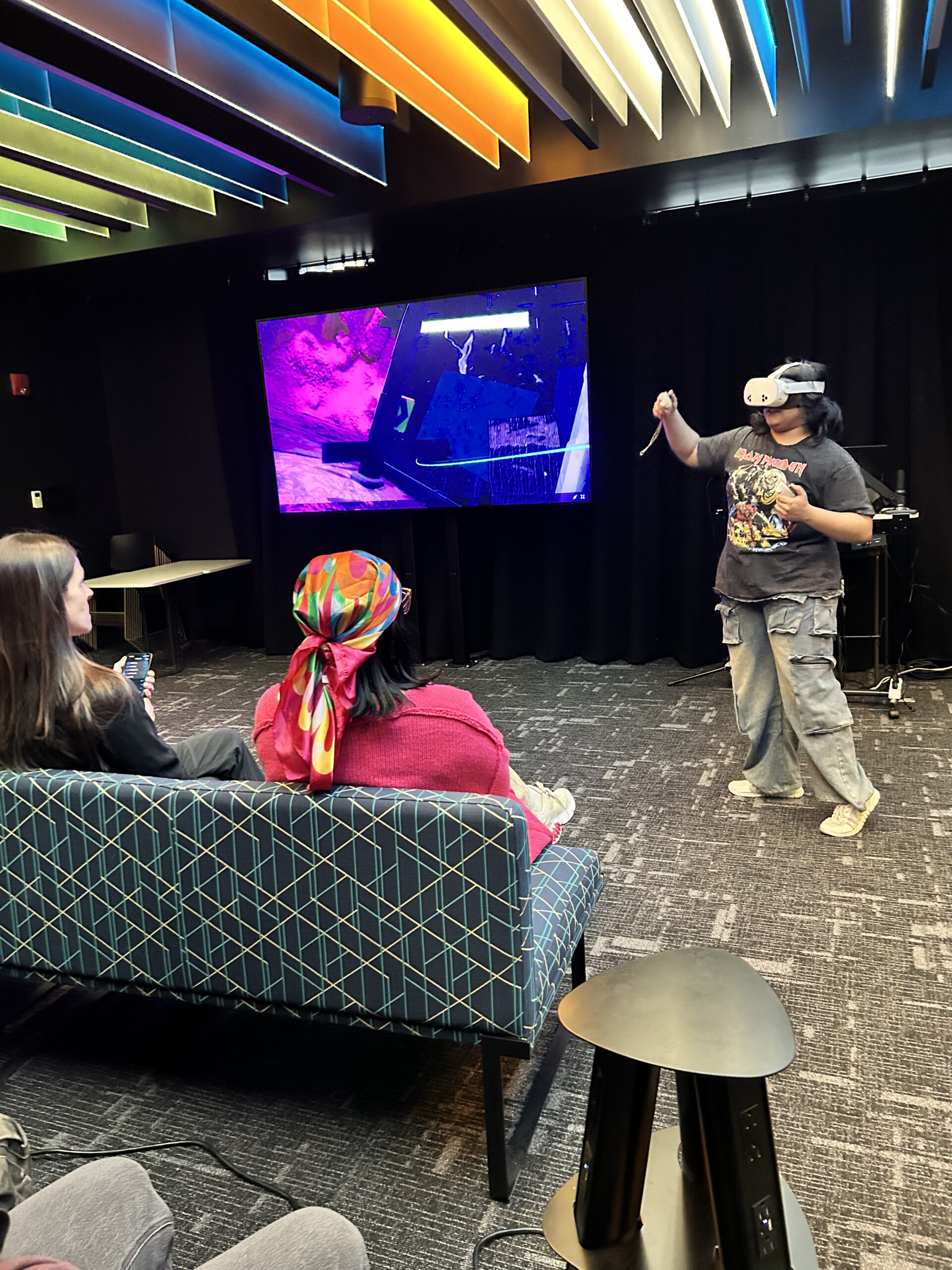 Person wearing a virtual reality headset and holding motion controllers demonstrates an immersive VR experience in a presentation space while seated attendees observe. A large display screen mirrors the virtual environment, and colorful illuminated ceiling panels brighten the room as audience members watch the interactive presentation from nearby couches.