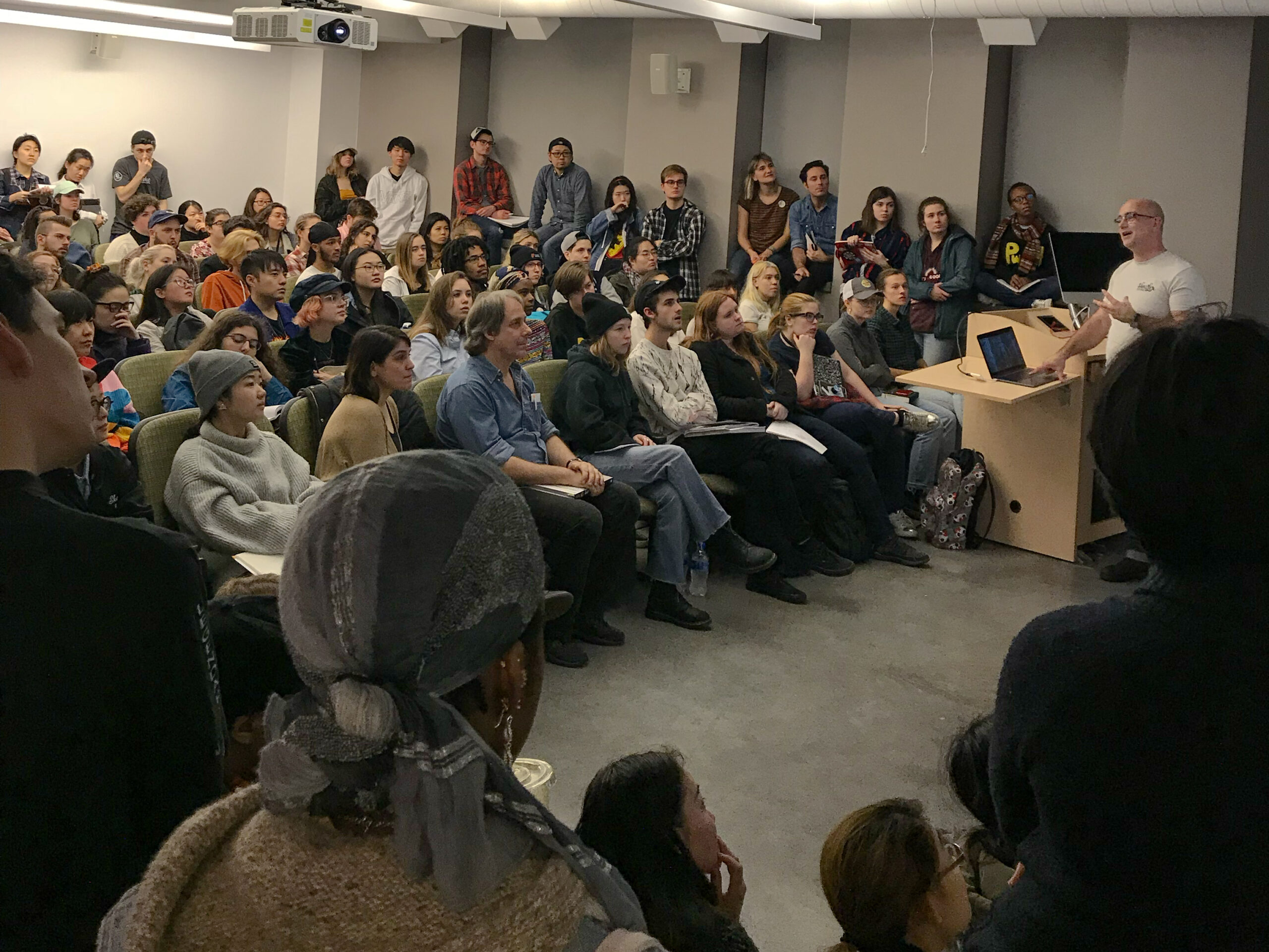 A guest speaker addresses a packed classroom of students seated and standing throughout the room during a lecture. Students watch attentively while the presenter speaks beside a podium with a laptop, and audience members hold notebooks and sketchbooks for note-taking.