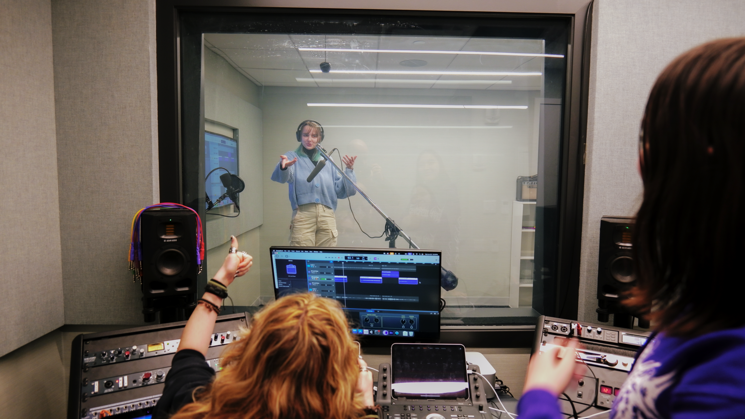 Student records vocals in a professional soundproof recording booth while another student operates the control room workstation using digital audio workstation software and studio mixing equipment. Through the soundproof glass, the performer gestures while speaking into a studio microphone wearing headphones as the recording engineer monitors the session using studio speakers, audio processors, and recording controls.