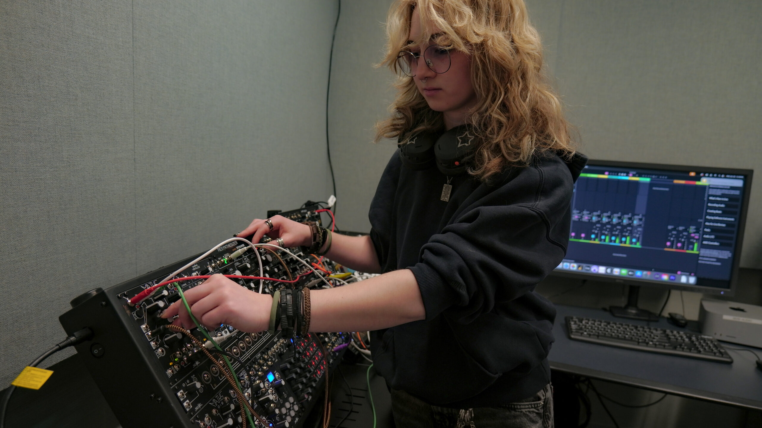 A student adjusts controls and patch cables on a modular synthesizer system in an electronic music production studio, with a computer displaying digital audio workstation software and studio monitor speakers positioned at the workstation.