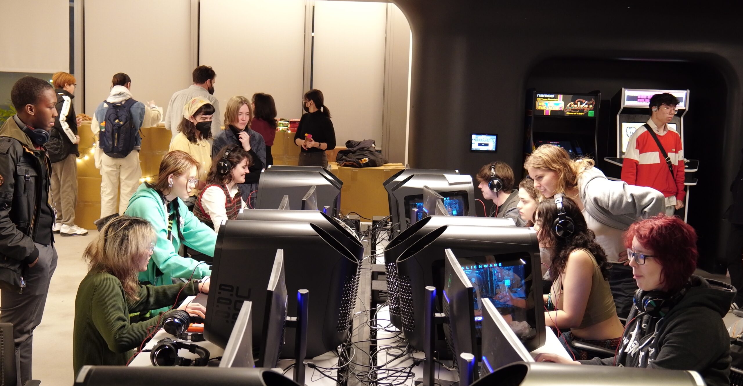 Students participate in a collaborative tabletop game design activity in a digital media lab, discussing game mechanics while holding printed game materials around a shared table. Desktop gaming computers with illuminated towers, monitors, keyboards, and arcade cabinets are visible throughout the space as other students work and play games in the background.