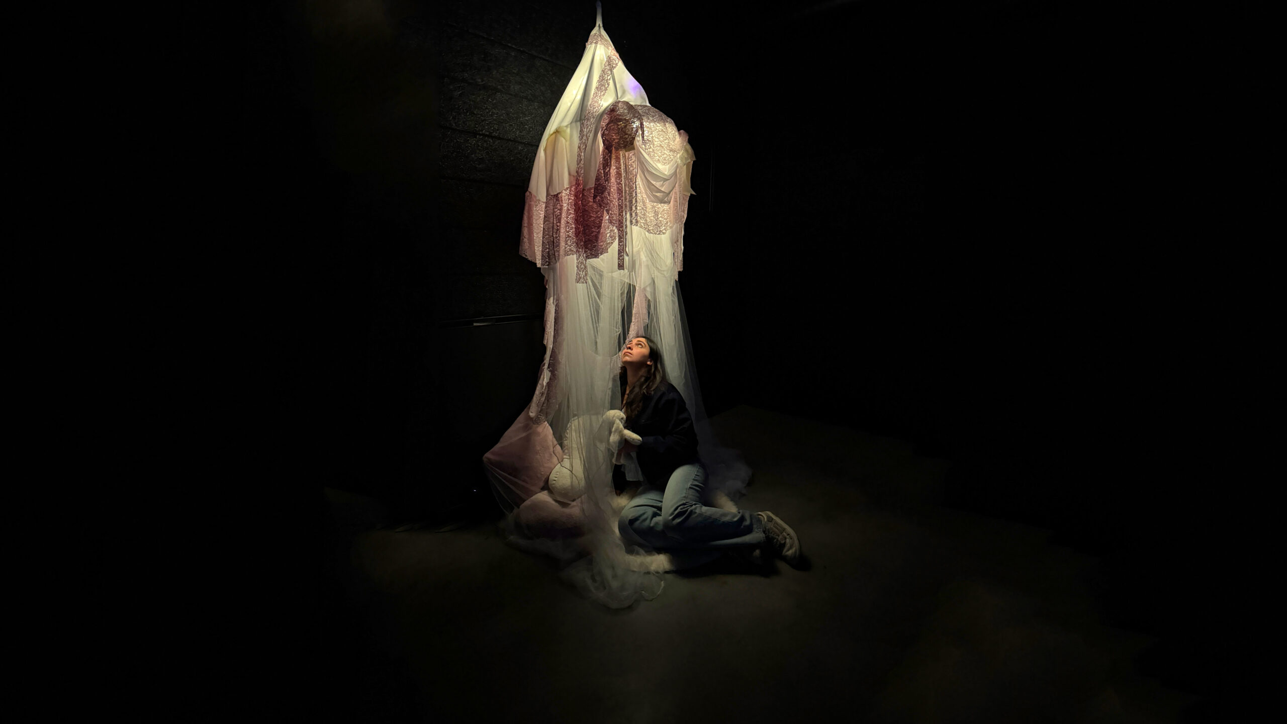 A person kneels beneath a suspended sculptural installation made of layered translucent fabric and lace in a dark gallery space, looking upward while holding a white plush object as soft spotlighting illuminates the artwork.