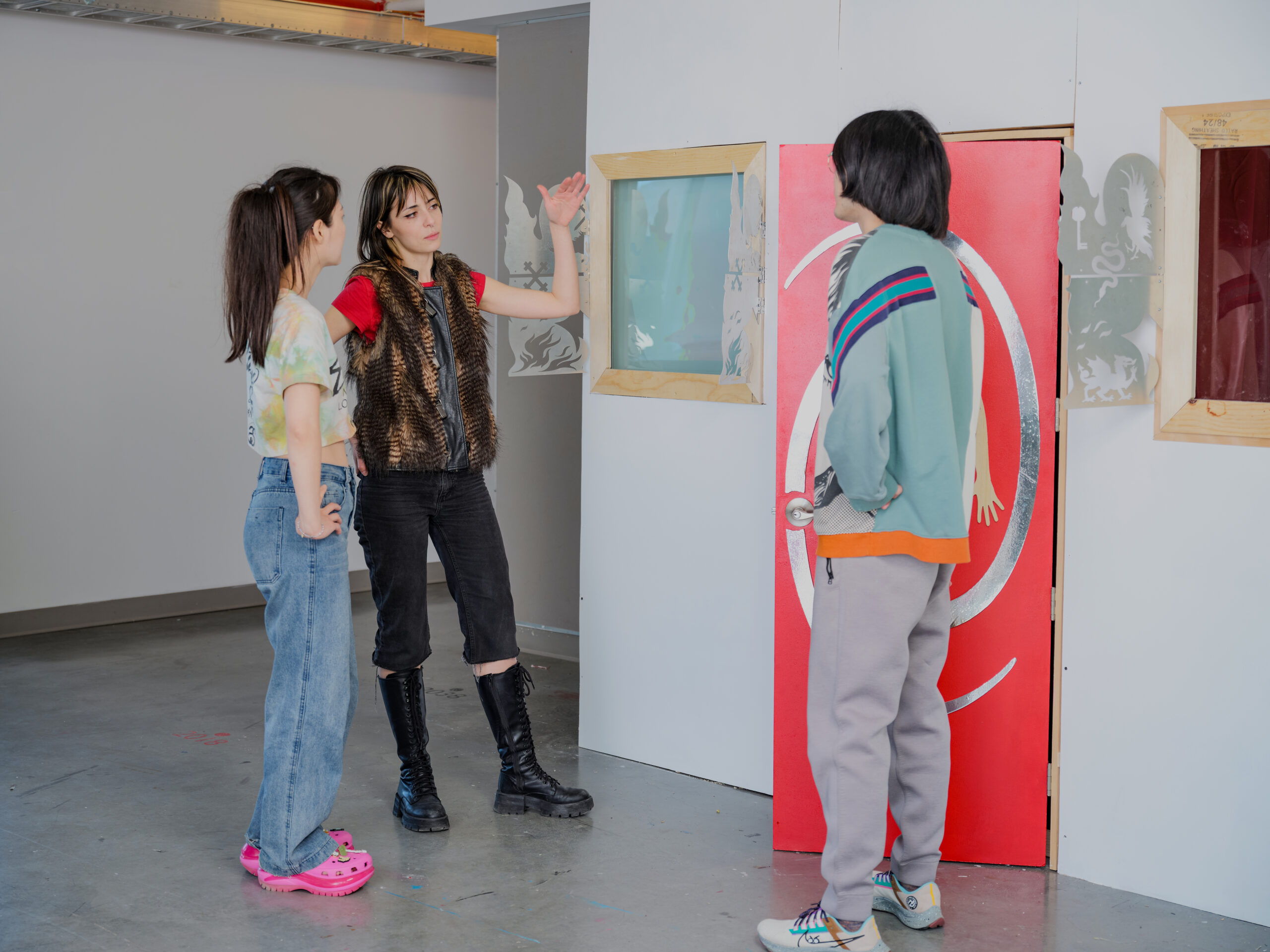 Three students stand in discussion beside an interactive installation featuring a bright red door, framed windows, and decorative cutout graphics. One student gestures while explaining the work as the others listen and examine the installation in the studio.