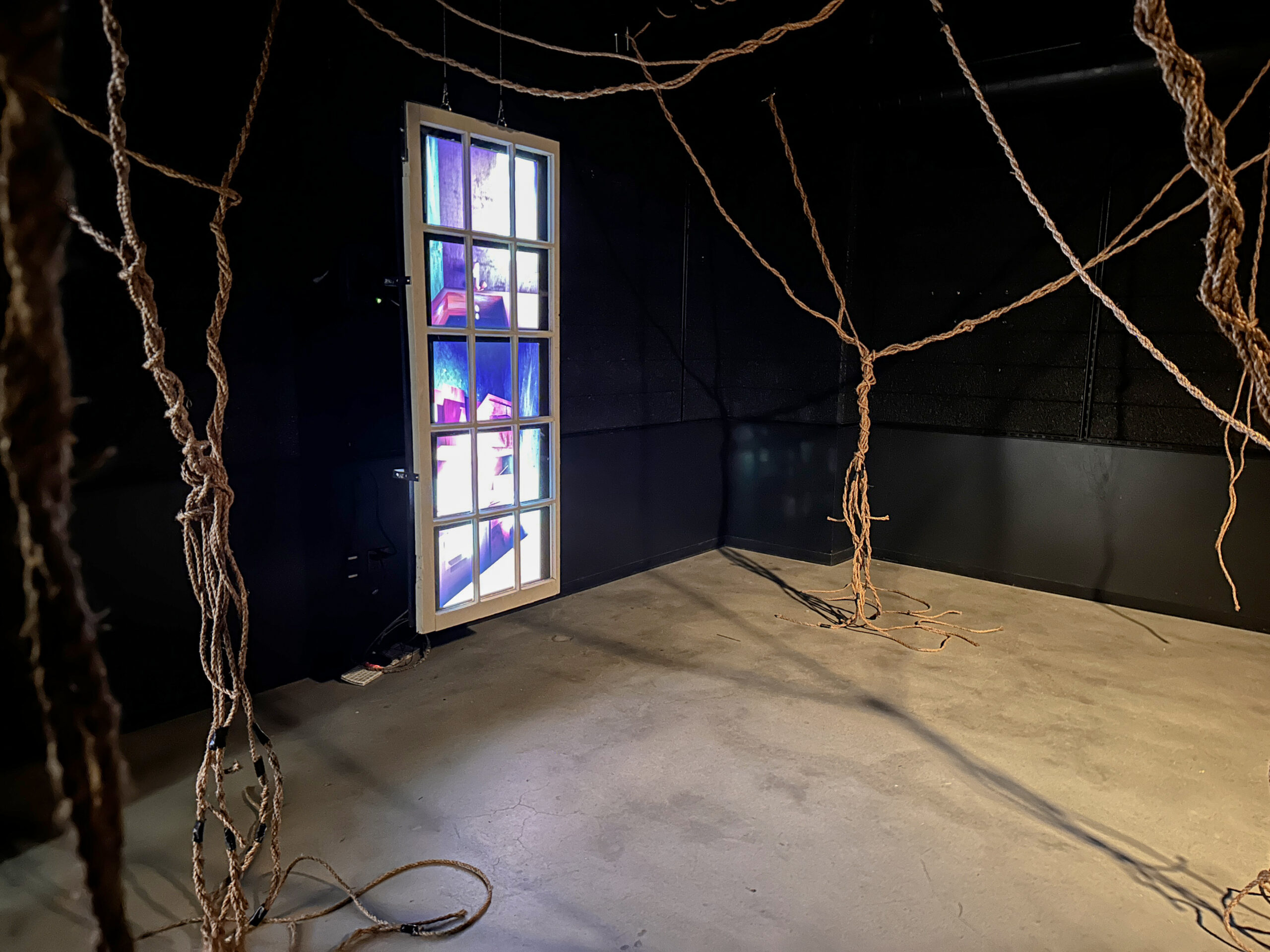 An immersive installation featuring suspended rope-like strands hanging throughout a dark gallery space, with a freestanding illuminated window displaying colorful moving imagery against black walls and a concrete floor.