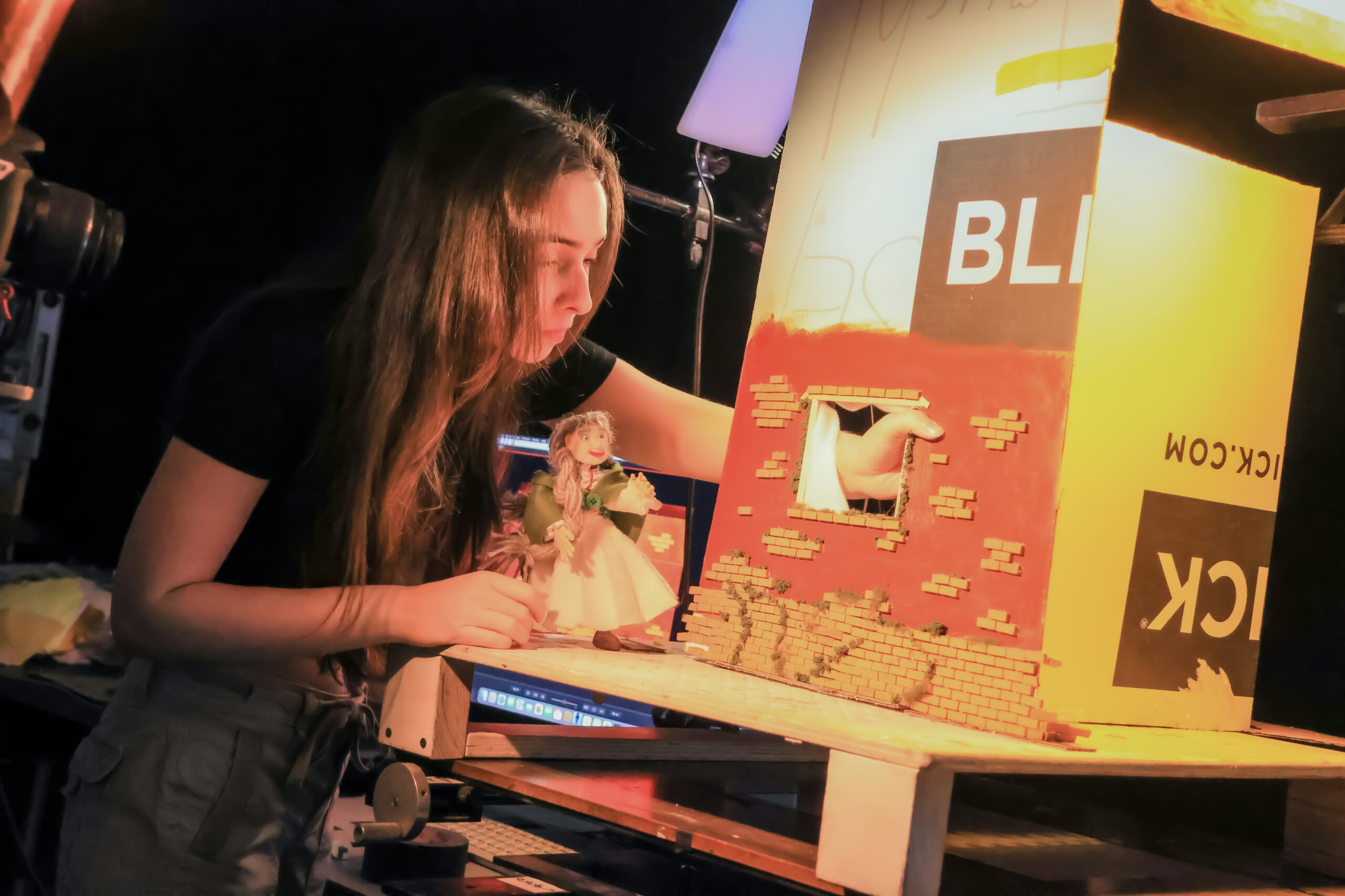 A student carefully positions a handmade puppet on a miniature stop-motion film set depicting a brick building façade with a window, while a DSLR camera and studio lighting are arranged to capture the animated scene.