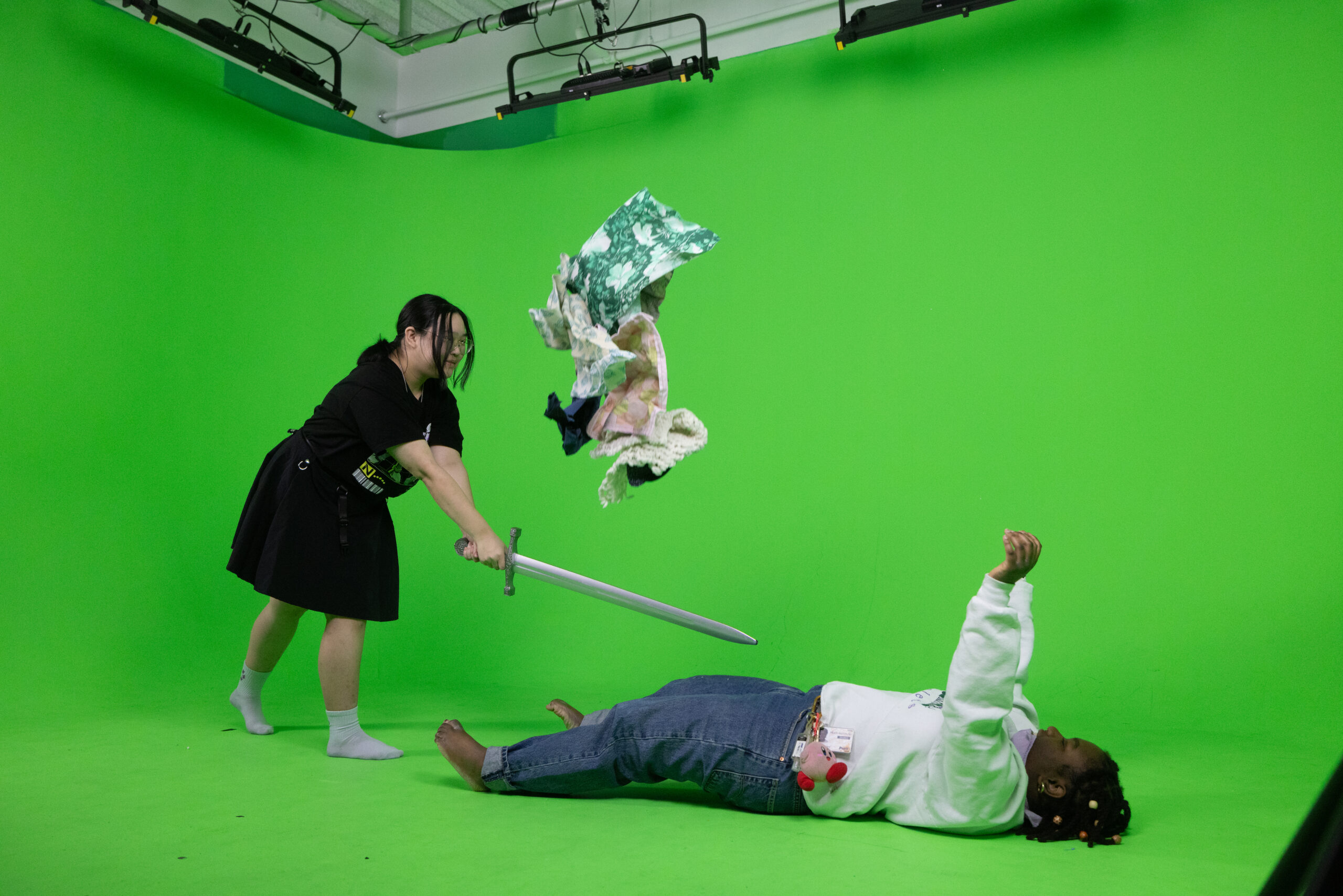 A student in a green screen studio swings a prop sword toward another student lying on the floor while pieces of fabric are suspended in midair, demonstrating a staged visual effects scene for film or animation production beneath overhead studio lighting.