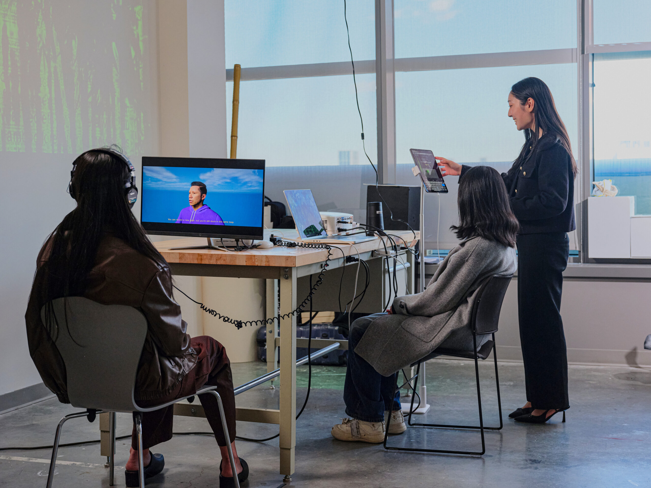 A student presents a digital media project while standing beside a table with a laptop, monitor, speakers, and tablet. Two audience members wearing headphones sit facing the presentation as a video with subtitles plays on the monitor and abstract imagery is projected onto the wall.