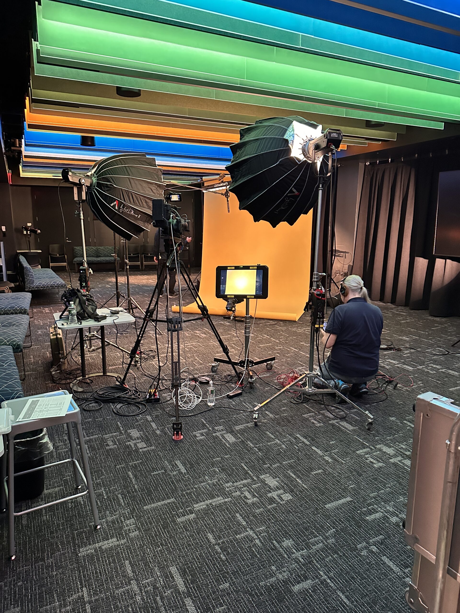 Professional video production setup in a multipurpose presentation space with large softbox studio lights, cameras on tripods, monitors, cables, and an orange seamless backdrop. A crew member adjusts equipment while colorful illuminated ceiling panels span the room, which has been converted into a temporary filming studio.