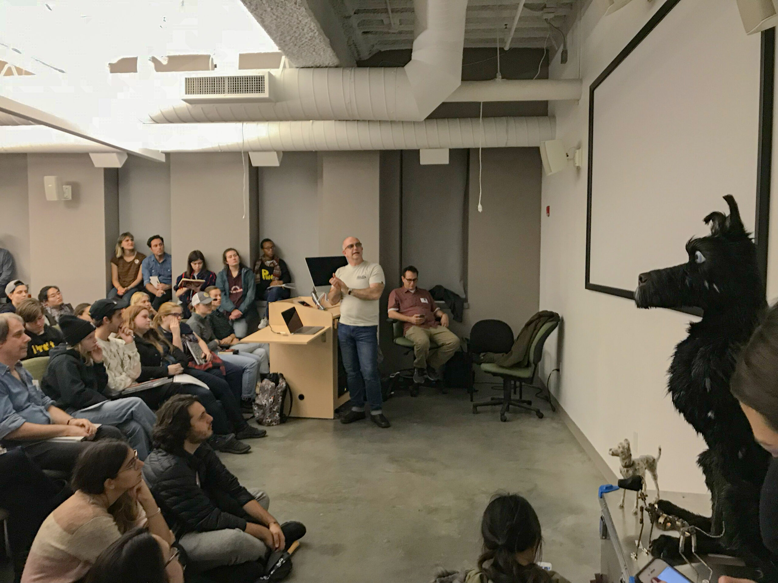 A guest speaker presents to a full classroom of students in a lecture space while standing beside a podium and projection screen. A large black puppet resembling a dog and smaller articulated animal puppets are displayed at the front of the room as demonstration pieces.