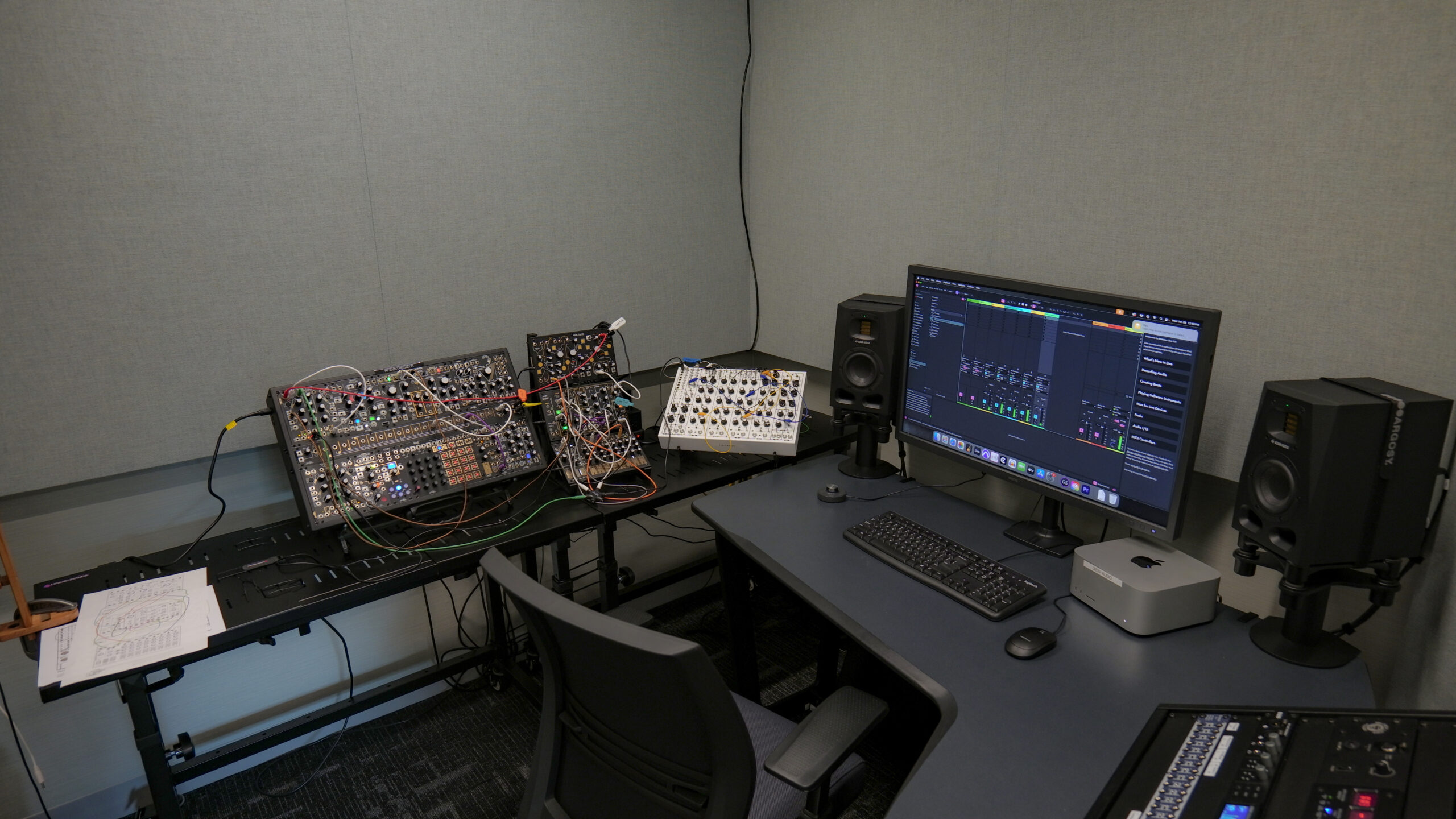 An electronic music production studio featuring modular synthesizer systems connected with patch cables, a desktop computer running digital audio workstation software, studio monitor speakers, a keyboard, and audio production equipment arranged for sound design and music composition.