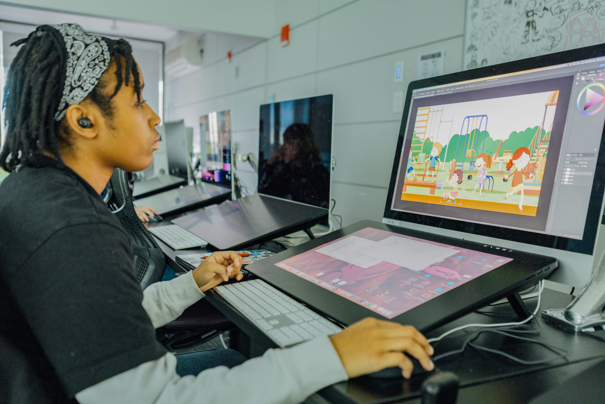 A student works at a digital drawing workstation using a pen display while creating a colorful illustration of children playing on a playground. Additional drawing tablets and computer workstations are visible throughout the animation studio.
