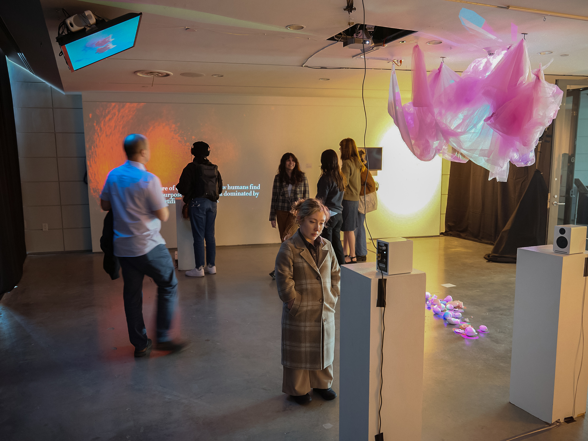 Three students stand together using a desktop computer at an interactive digital art installation. One student operates the keyboard and mouse while the others watch the screen, one holding a takeaway coffee cup. A white speaker sits on a pedestal in the foreground, and a glass entrance with people visible outside is in the background.